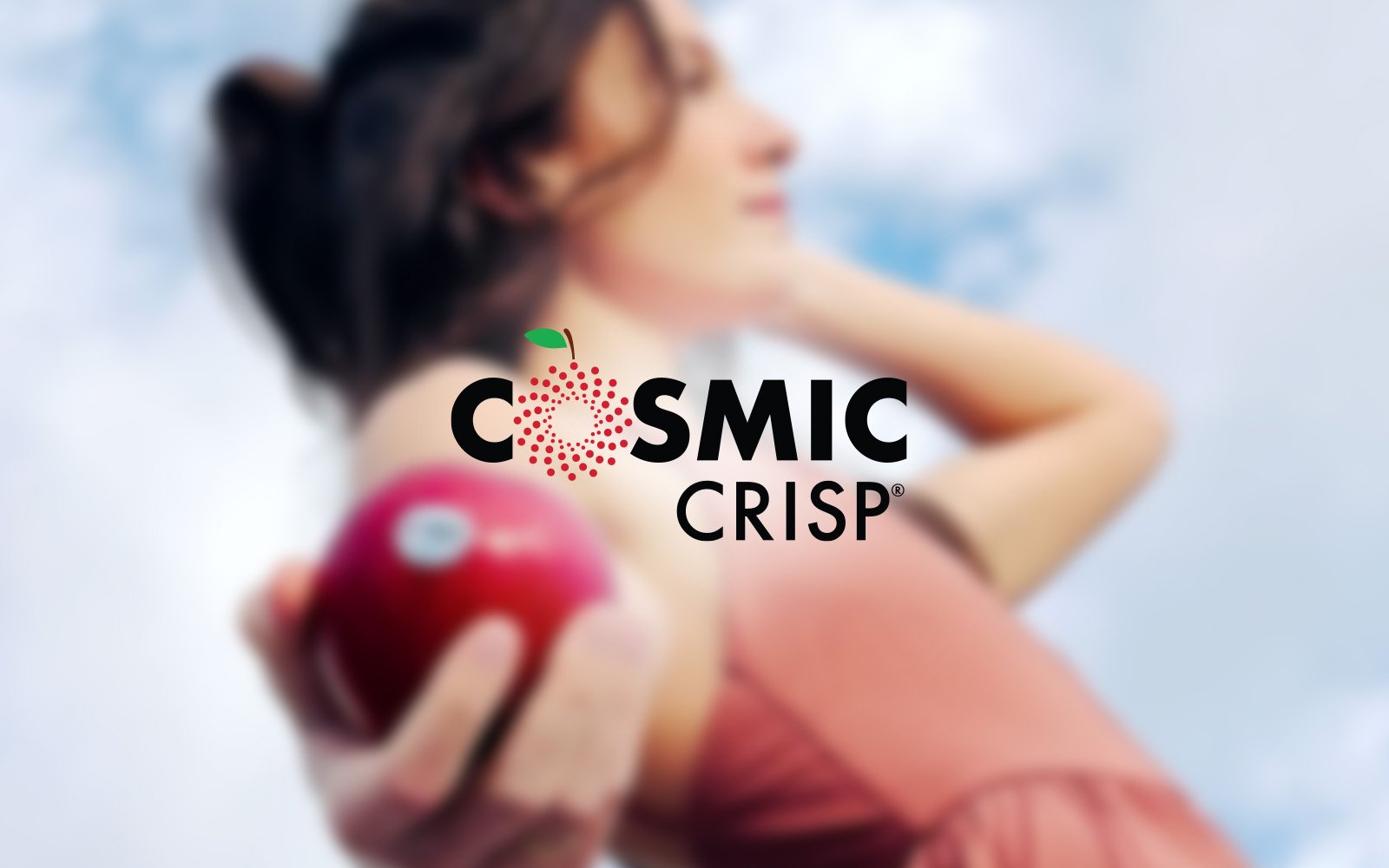 A woman with brown hair is holding a red apple towards the camera, with a blue sky in the background. The logo 'COSMIC CRISP' is overlaid on the image.
