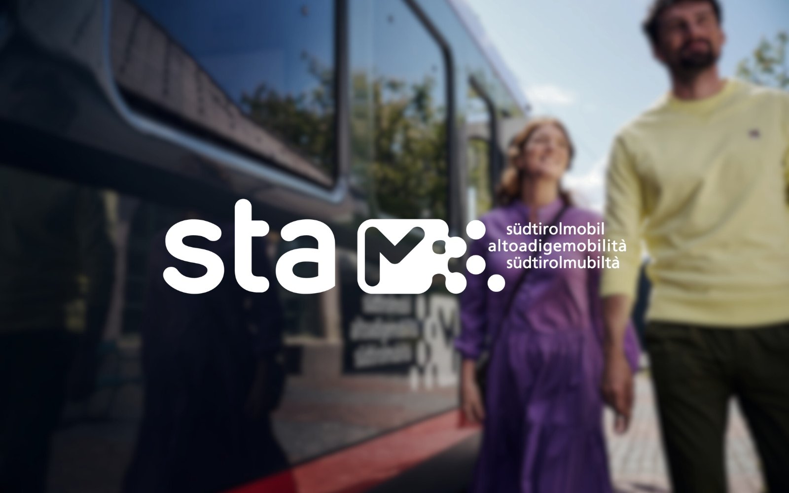 Two people standing outdoors near a bus, with their reflection visible in the bus window, smiling and enjoying a sunny day. With the name of the brand "STA" and its logo in the foreground.