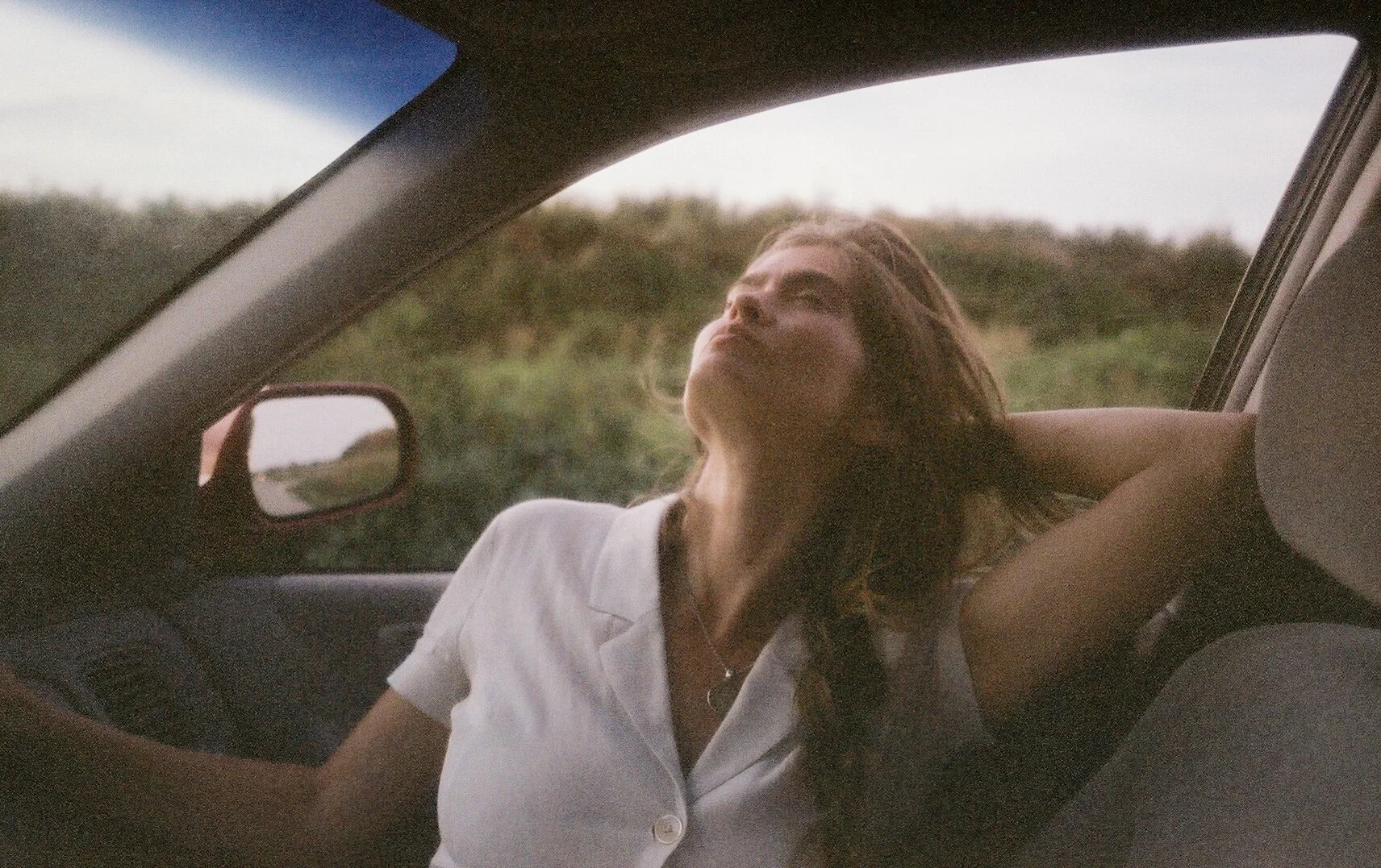 Young woman with long hair relaxing in the passenger seat of a car with her arm behind her head and eyes closed, outdoors in a natural setting.