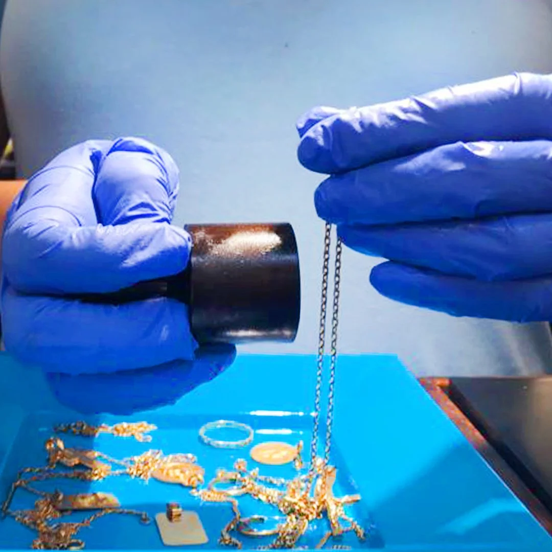 Manos con guantes de látex sosteniendo un recipiente con polvo blanco y una cadena, sobre una mesa con joyas y objetos decorativos.