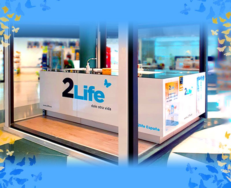 Mostrador de una tienda llamada '2Life' con logotipo de una mariposa, en un centro comercial, con señalización y productos visibles en el fondo.