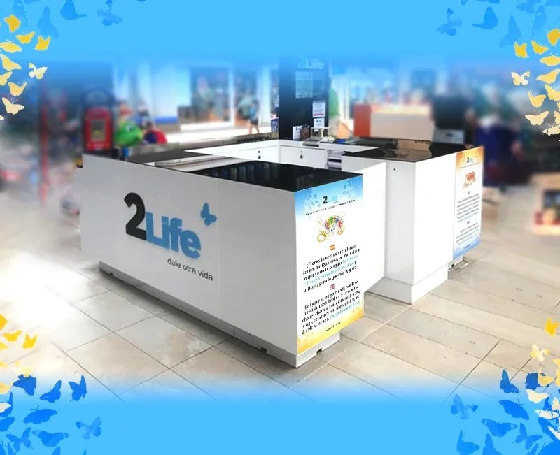 Mostrador de donaciones en un centro comercial con el logotipo 2Life y frases en español, rodeado de mariposas en los bordes de la imagen.