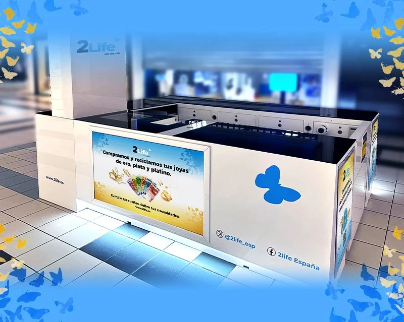 Un puesto de reciclaje de joyas de oro, plata y platino en un centro comercial, con carteles informativos y decoración de mariposas azules y amarillas.