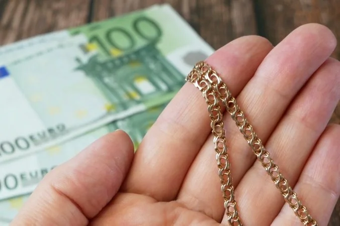 Una mano sosteniendo una cadena dorada y un billete de cien euros en un fondo de madera.