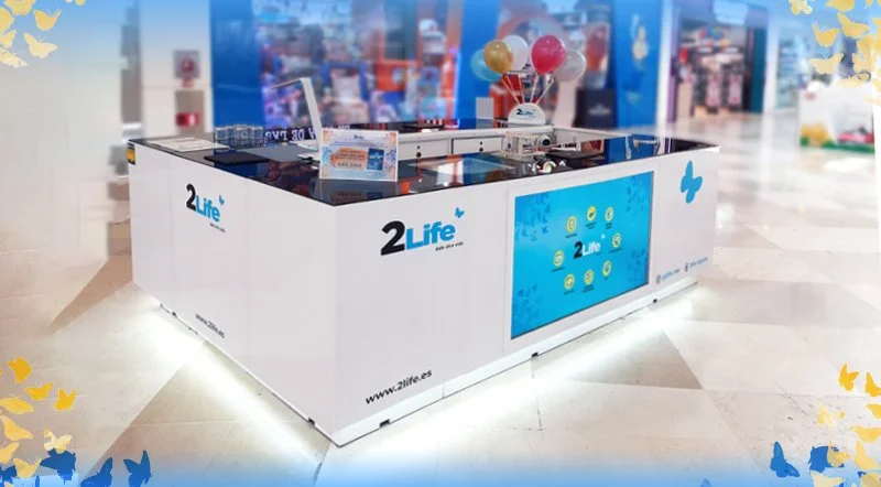 Estación interactiva de 2Life en un centro comercial con pantalla digital y globos de colores en la parte superior.