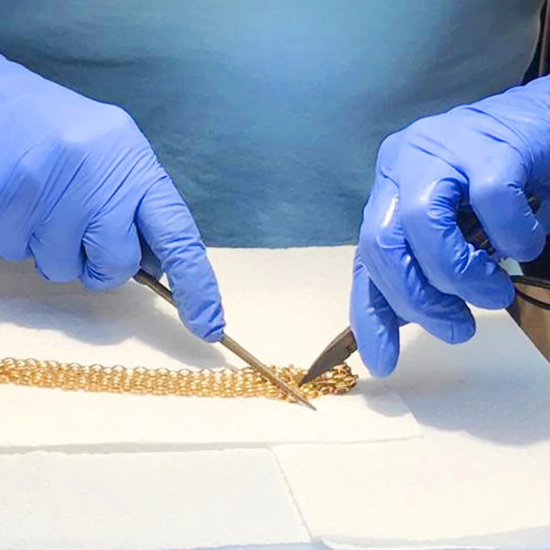 Manos con guantes azules manipulando una cadena de oro con pinzas.