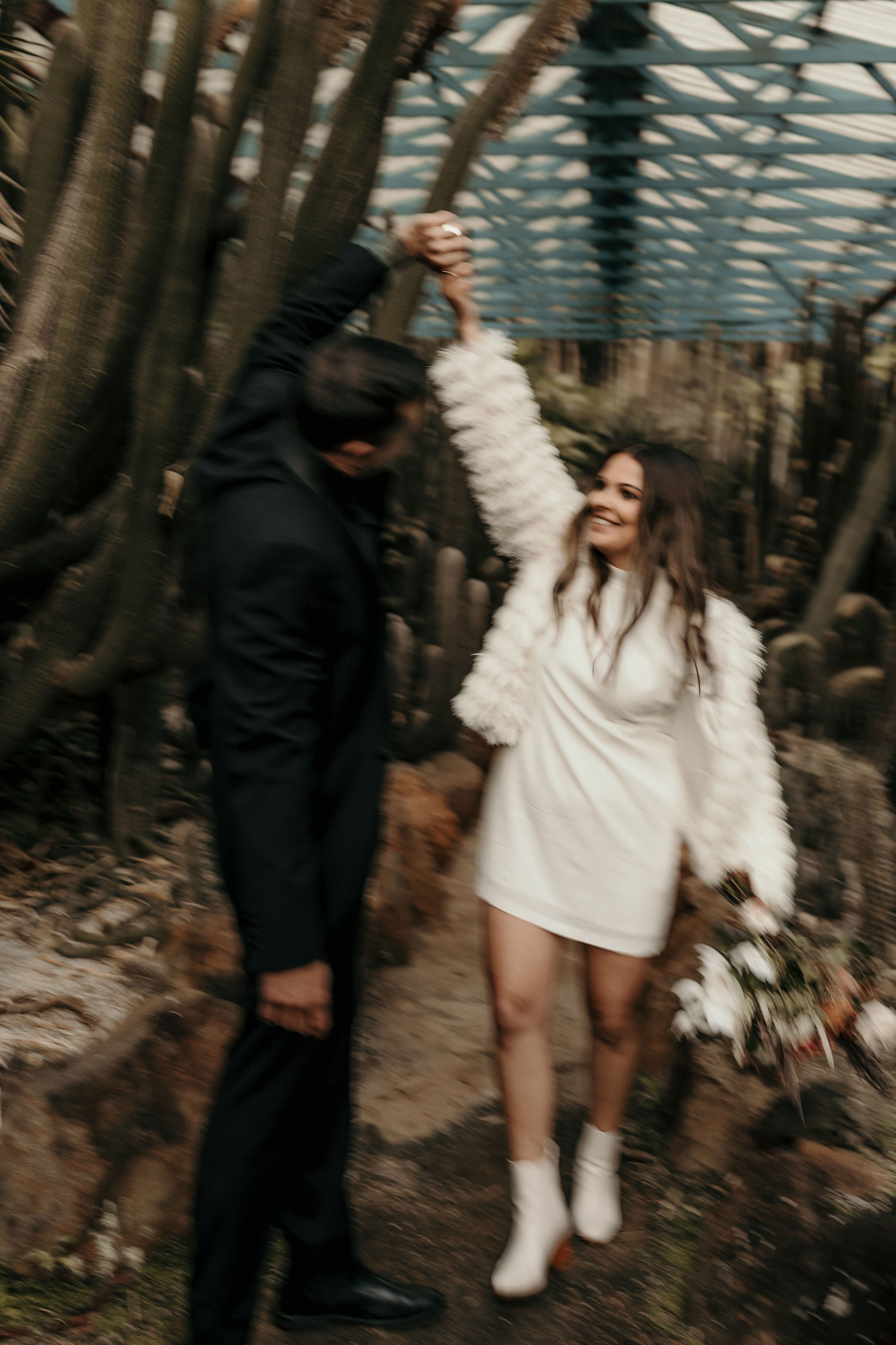 A couple dancing together outdoors in a forested area, with one person holding the other's hand up in the air. The woman is wearing a white dress and white boots, holding a bouquet of flowers, and smiling. The man is dressed in dark clothing, with hi