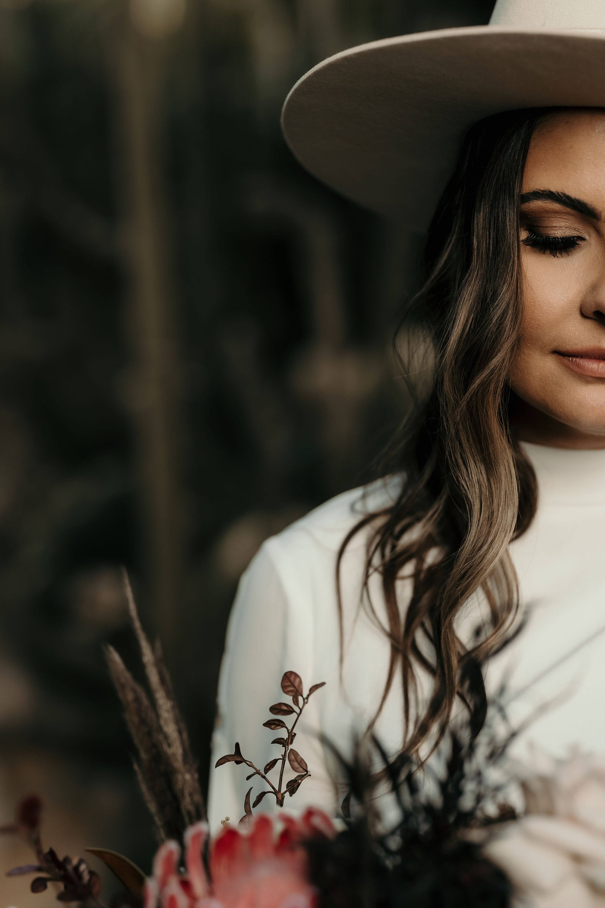 A woman wearing a beige wide-brimmed hat and a white dress, with long wavy brown hair, standing outdoors with blurred trees and plant life in the background.