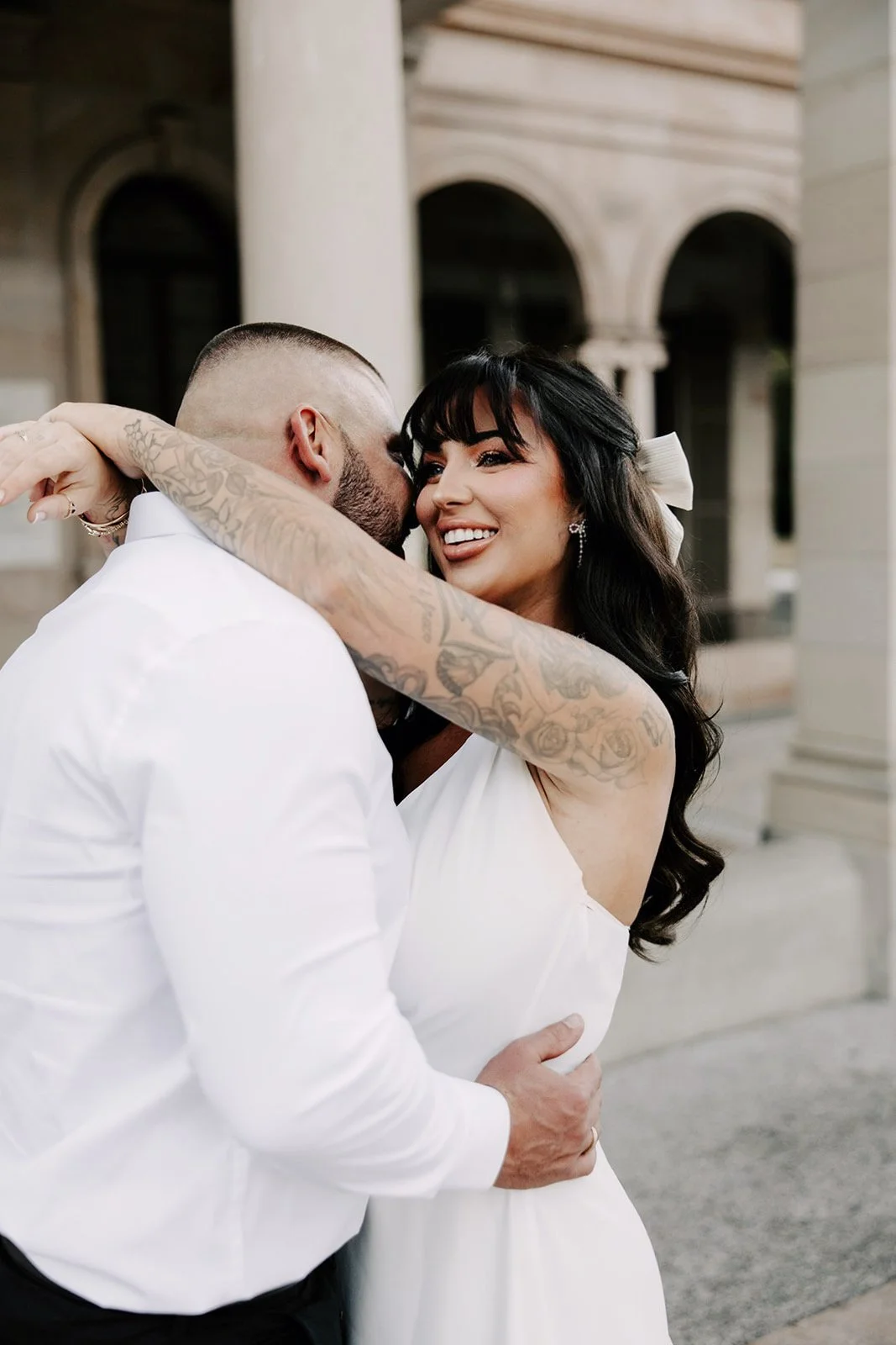 A couple in wedding attire sharing a close embrace outside a stone building. The woman has dark hair with a white bow and tattoos on her arm, smiling as she kisses the man. The man has a buzz cut and a beard, holding her waist.