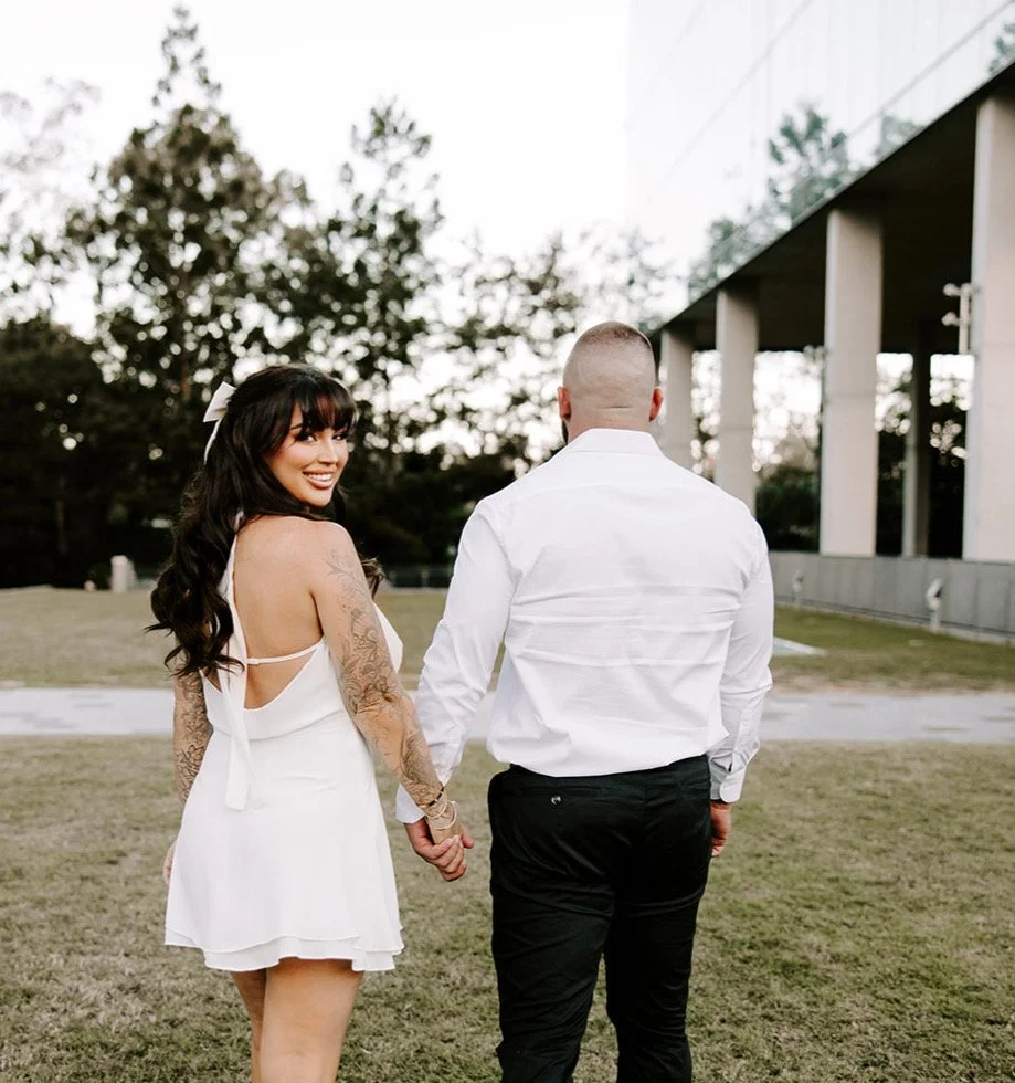 A woman with long dark hair, tattoos on her arms, wearing a white dress, holding hands with a man with a shaved head, wearing a white shirt and black pants, standing outdoors on grass near trees and a modern building.