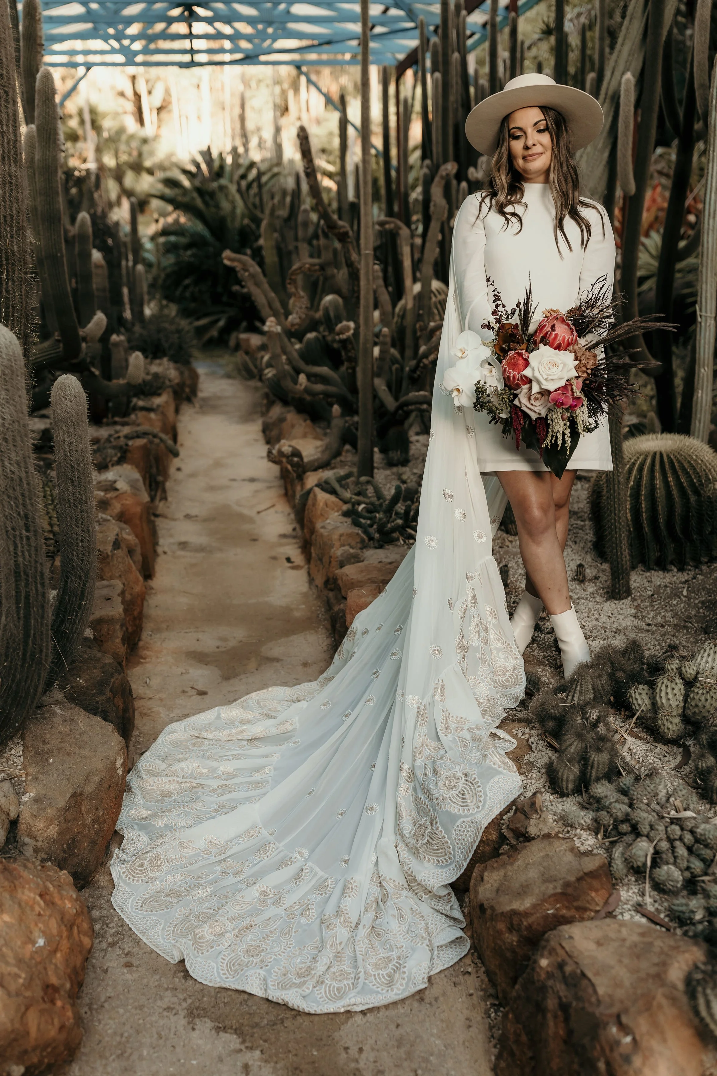 Woman in a white dress and wide-brimmed hat holding a bouquet of flowers in a desert plant greenhouse.