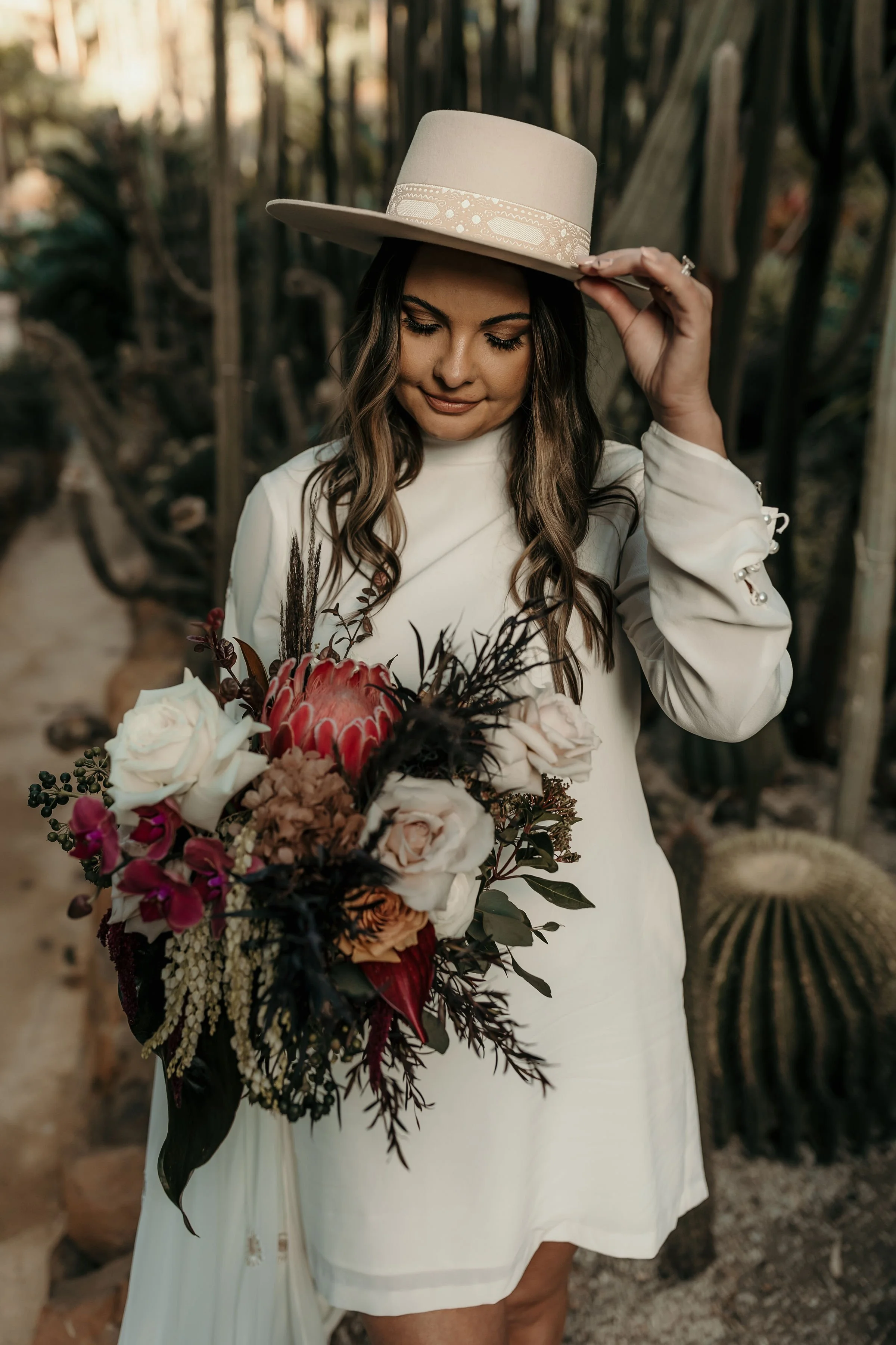 A woman wearing a white dress and a beige wide-brimmed hat is holding a large bouquet of flowers, standing outdoors among tall cacti and desert plants.