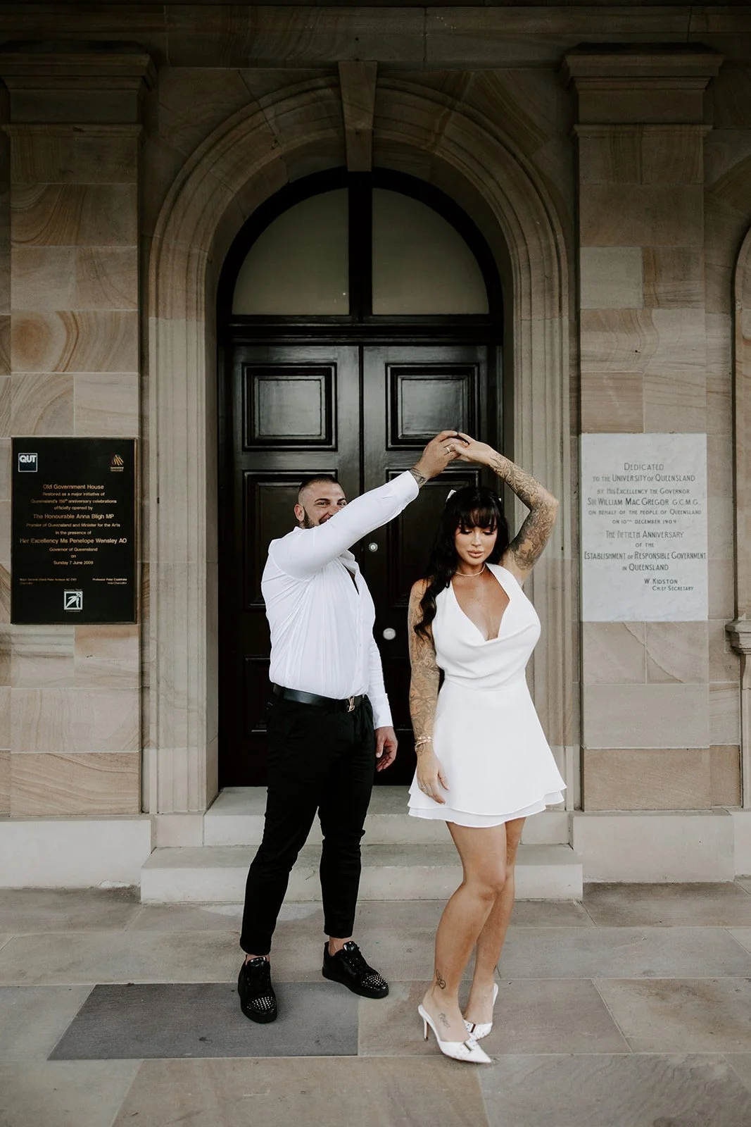 A couple dancing in front of a historic government building, the man is spinning the woman who wears a white dress, white high heels, and has tattoos, while the man wears black pants, black shoes, and a white shirt.