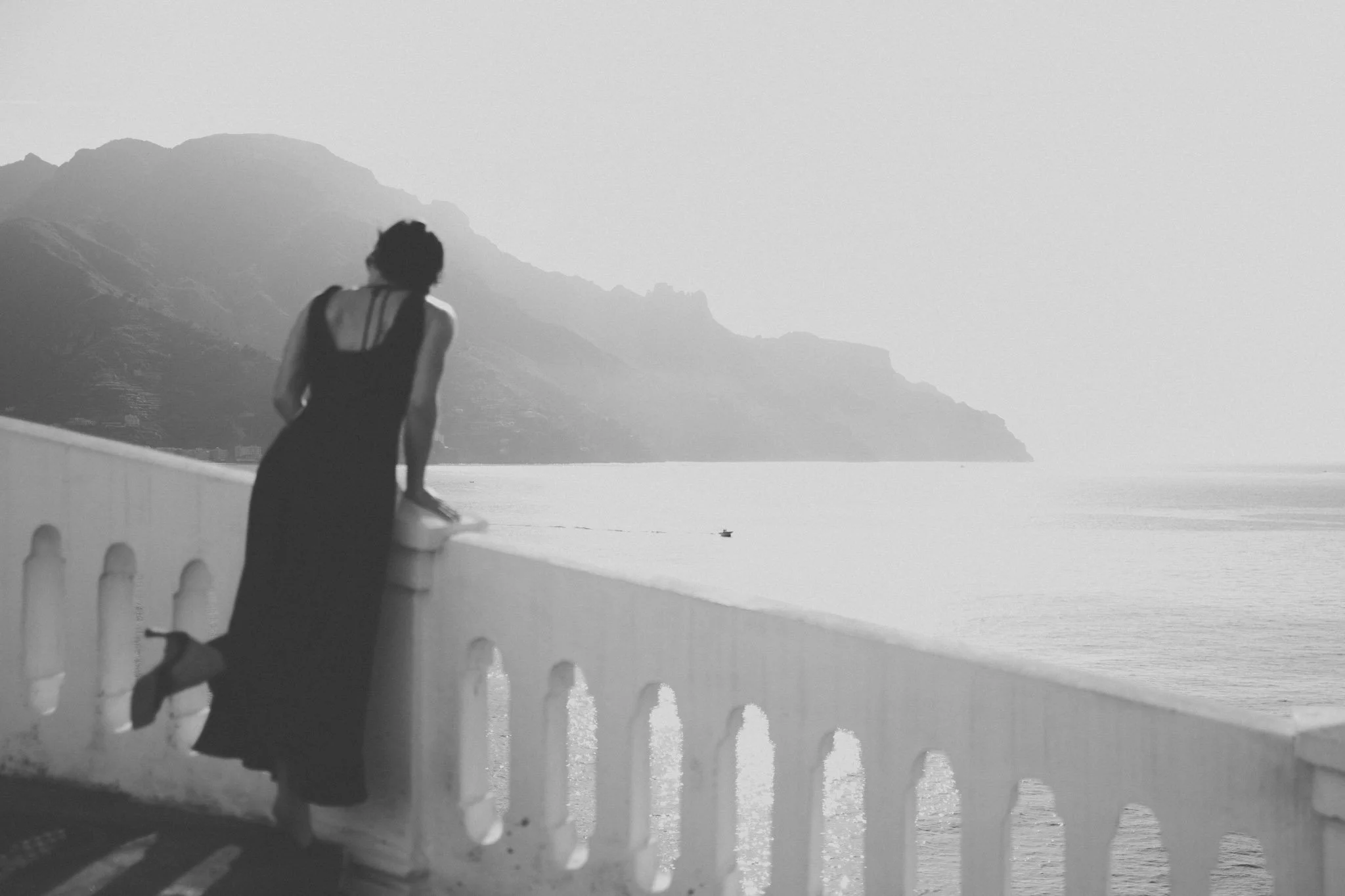 Eine Frau in einem langen Kleid lehnt an einer weißen Balustrade und schaut auf das Meer und die Berge im Hintergrund.