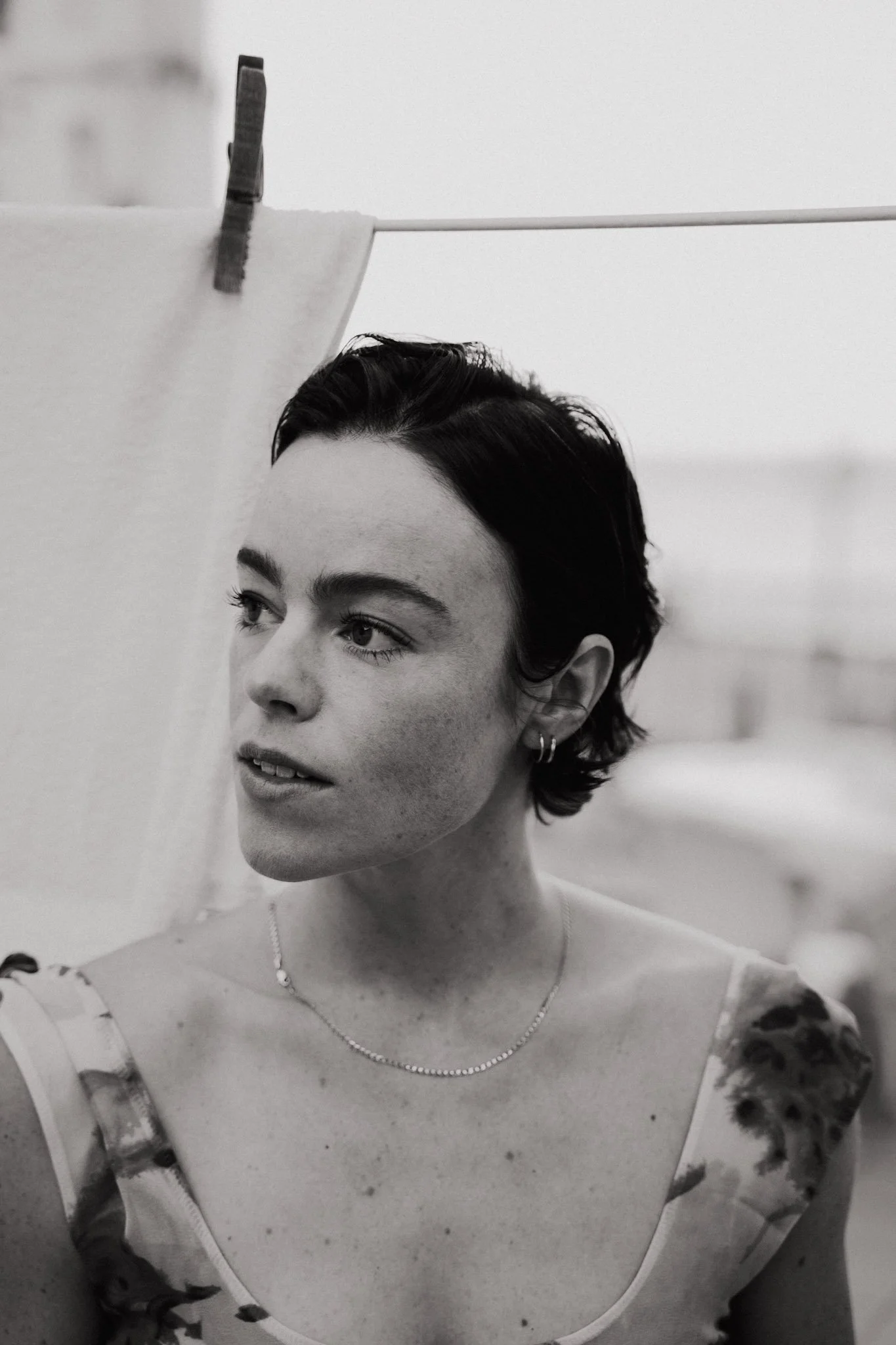 Schwarz-Weiß-Foto einer jungen Frau mit kurzen, dunklen Haaren, die an eine Wäscheleine mit einer Wäscheklammer hängt, trägt ein aufgeschnittenes Top und kleine Ohrringe, schaut leicht nach links, im Hintergrund unscharf.