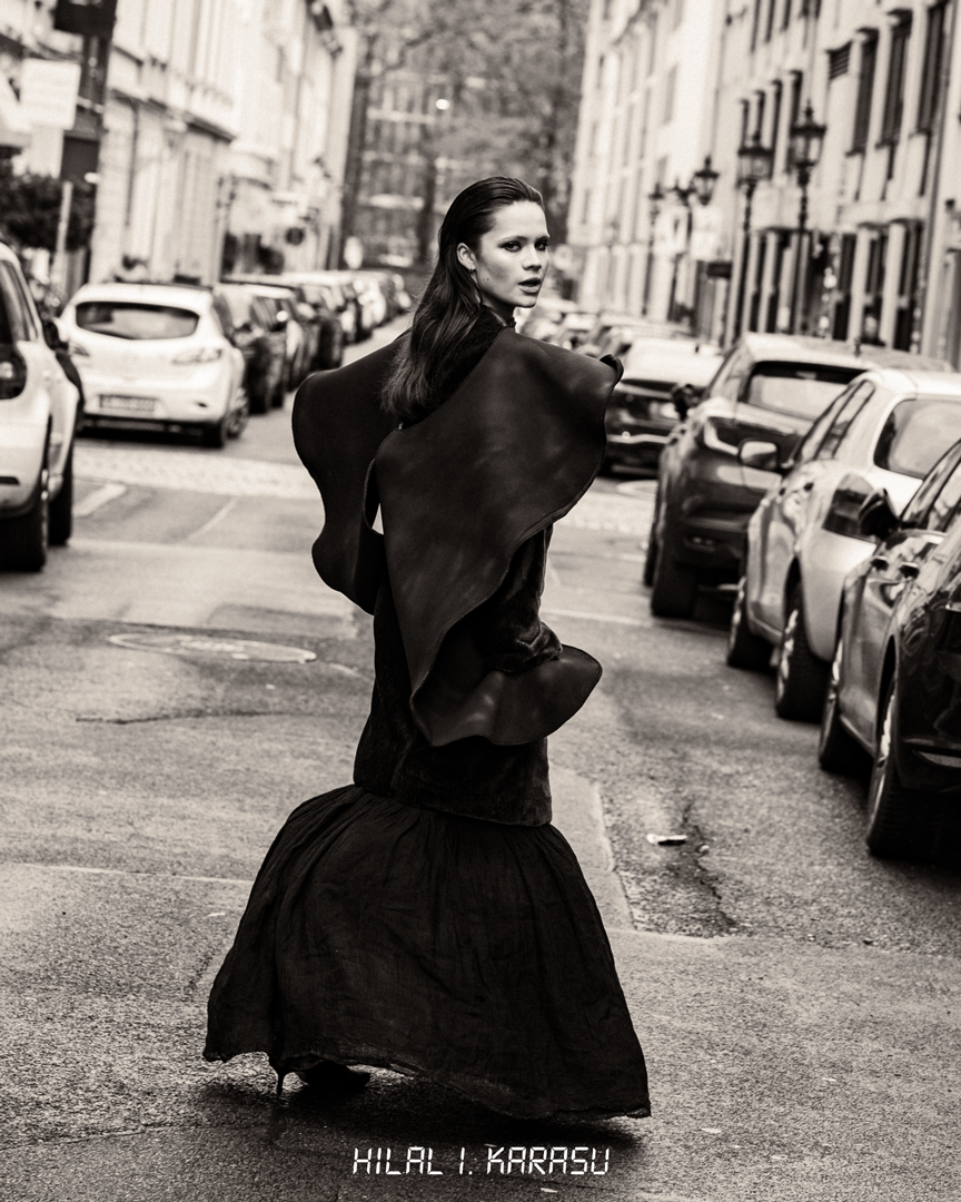 Schwarz-Weiß-Foto einer Frau, die auf einer Stadtstraße steht, mit Parkplätzen auf beiden Seiten, umgeben von Mehrfamilienhäusern. Die Frau trägt ein elegantes dunkles Kleid und einen großen schwarzen Schal.