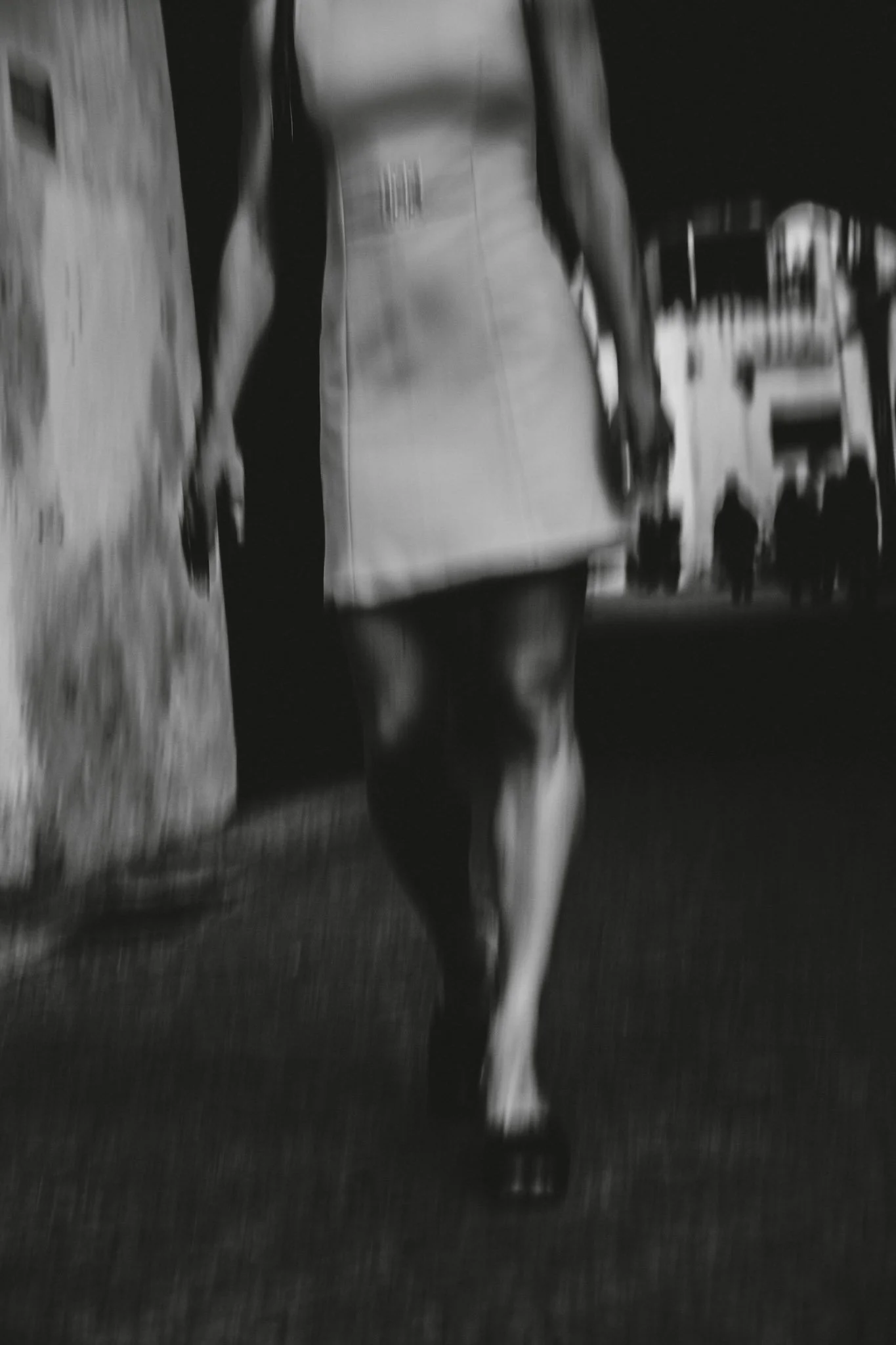 Schwarz-weiß-Foto einer Frau, der ein Cocktailkleid trägt, zu sehen von vorn, auf Höhe der Schultern abgeschnitten. auf einer Straße mit einigen Menschen im Hintergrund.