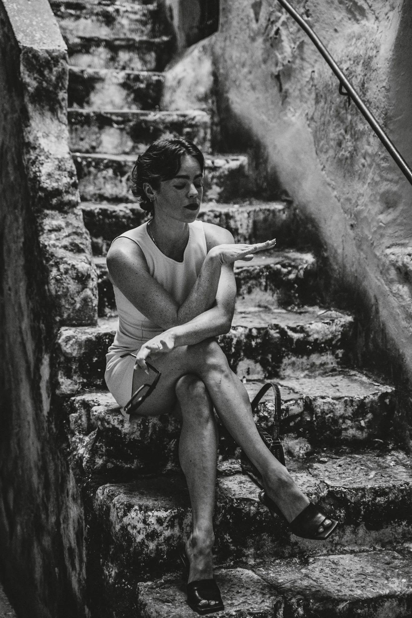 Schwarz-weiß-Fotografie einer Frau mit dunklen Haaren, sitzend auf alten Steinstufen, die Treppe hinunter, mit geschlossenen Augen, die Hand vor dem Gesicht hält und eine Sonnenbrille in der anderen Hand. Sie trägt ein ärmelloses Kleid und offene Sch
