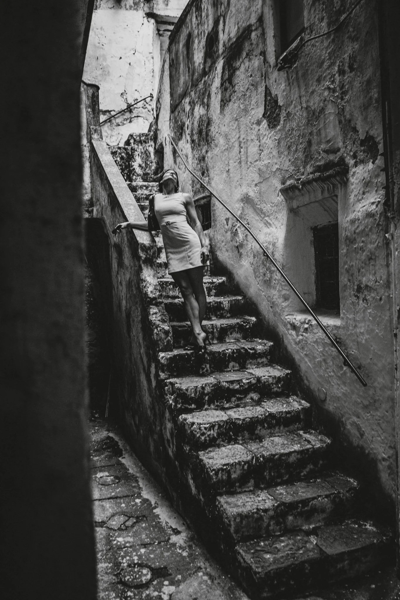 Schwarze und weiße Aufnahme einer Frau, die eine alte, steile, steinerne Treppe in einer engen, verfallenen Gasse hinaufgeht.