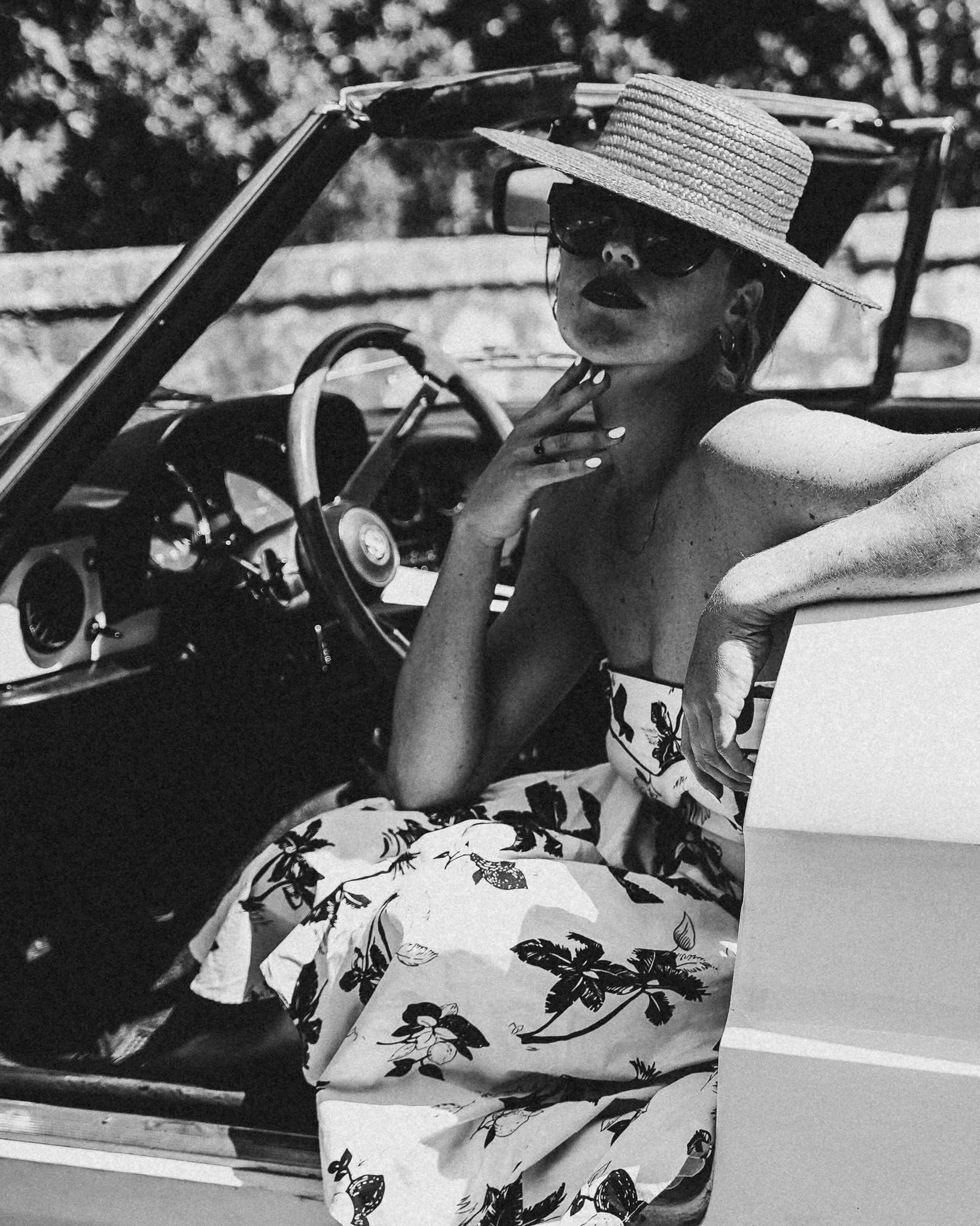 Schwarz-Weiß-Foto einer Frau in einem Cabriolet, trägt eine große Sonnenbrille, einen Strohhut und ein Kleid mit Blumenmuster, sitzend im Auto.