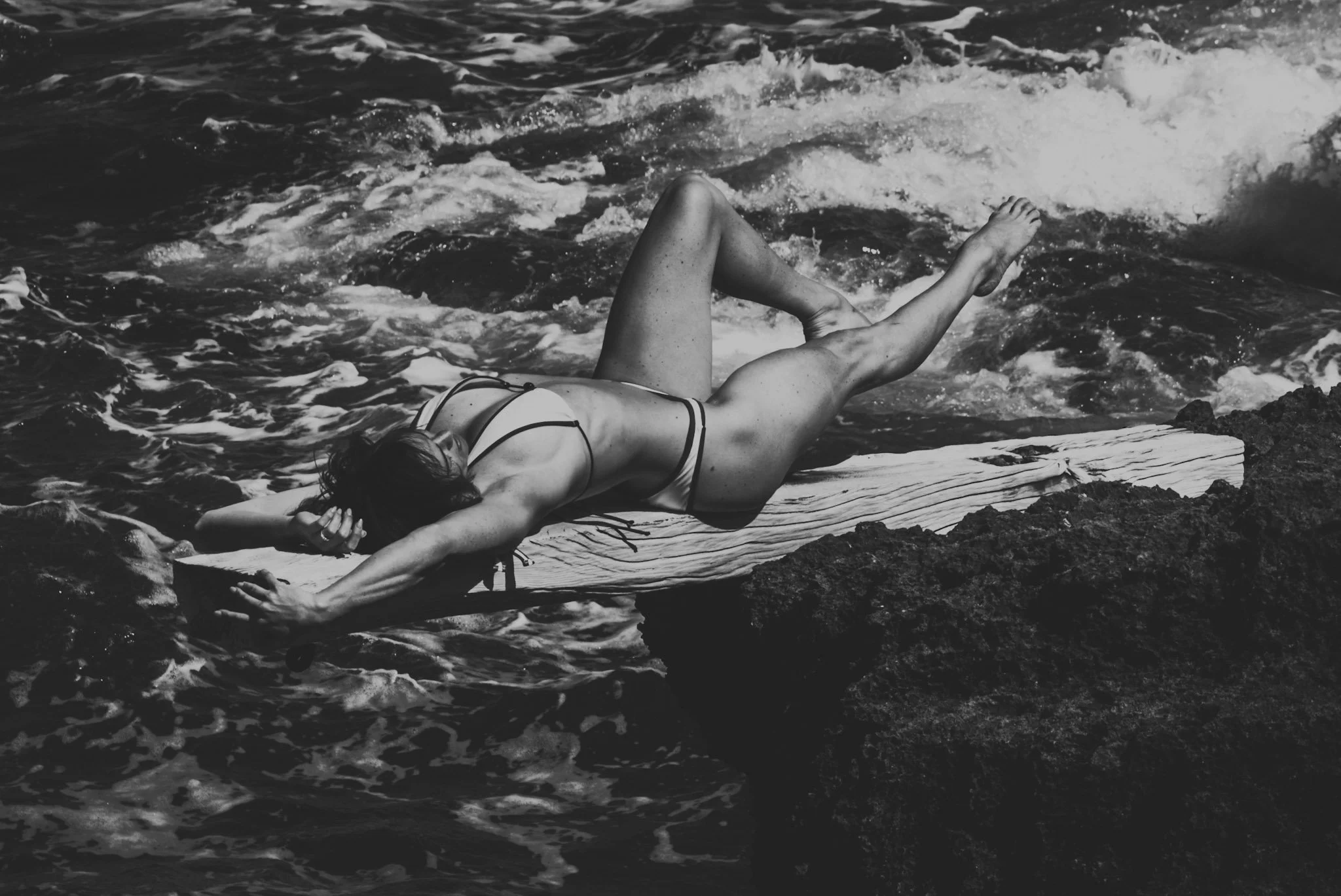 Schwarz-weiß-Foto einer Frau in Bikinis, die auf einem Surfbrett auf Felsen am Meer liegt, Wellen im Hintergrund.