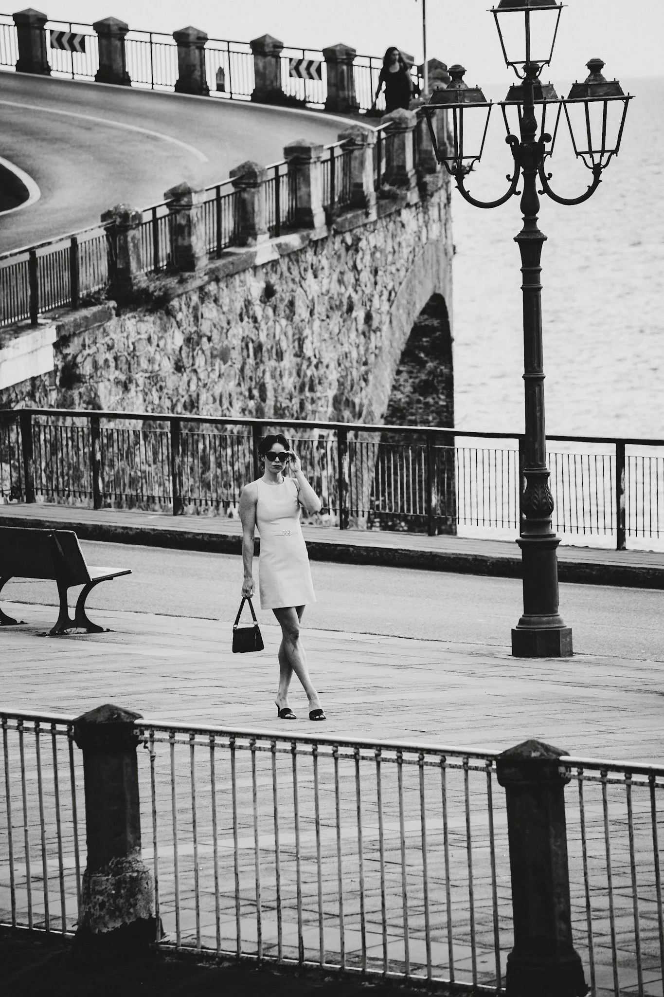 Eine schwarze und weiße Fotografie eines öffentlichen Platzes mit einer Frau in einem weißen Kleid, die eine Tasche trägt und eine Sonnenbrille trägt. Im Hintergrund eine Brücke über das Wasser und eine alte Straßenlaterne.