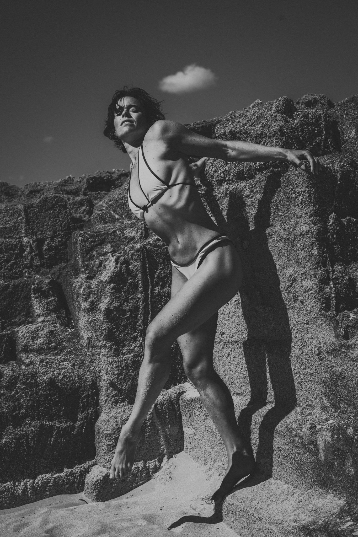 Schwarz-weiß-Fotografie einer Frau in Bademode, die an Felsen lehnt, mit einer kleinen Wolke im Himmel im Hintergrund.