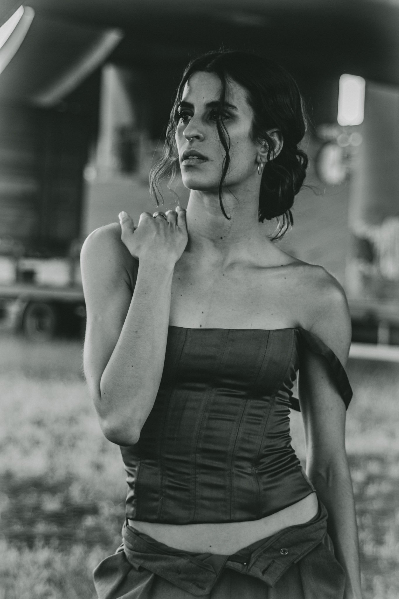 Schwarz-weiß-Fotografie einer jungen Frau mit dunklen, lockigen Haaren, die einen schwarzen, taillierten, trägerlosen Top trägt. Sie steht im Freien vor einem Lagerfeuer oder einer offenen Landschaft, mit leichten Unscharf-Hintergrund. Sie schaut nac