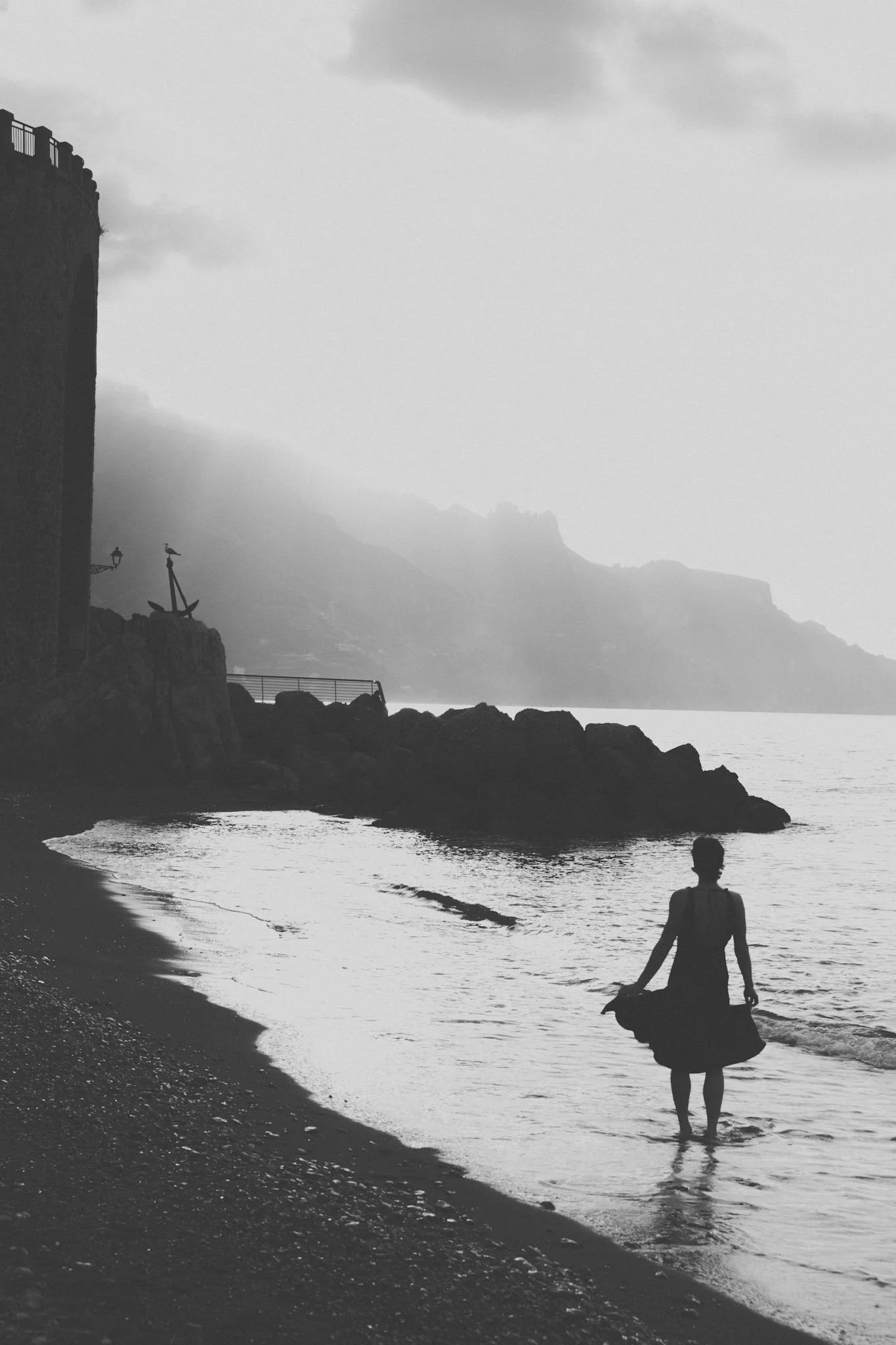 Schwarz-weiße Aufnahme einer einsamen Frau, die am Strand steht, mit Blick aufs Meer, bei bewölktem Himmel.