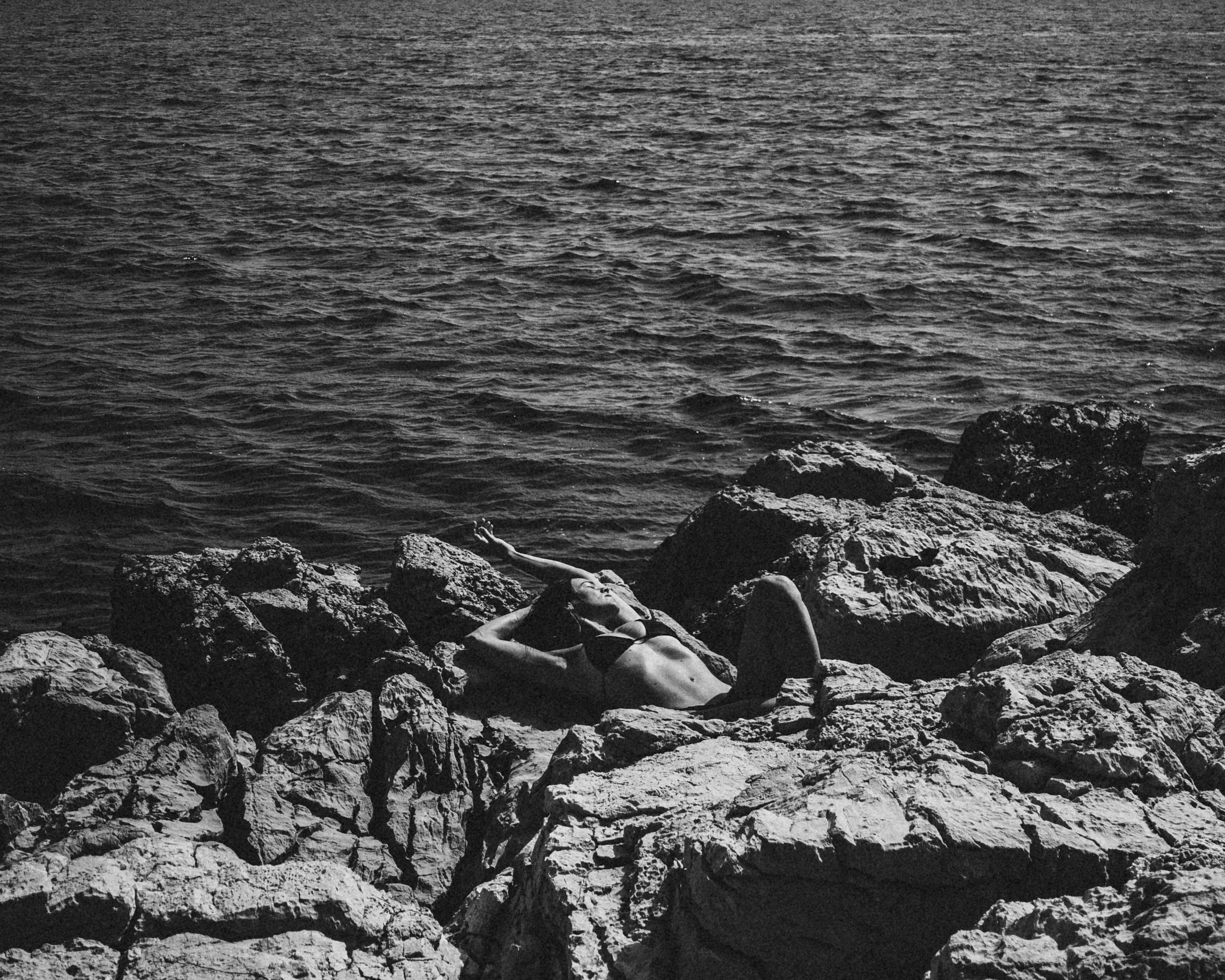 Frau liegt auf Felsen an der Küste bei Sonnenuntergang, Sonnenbrille, Bikini, Schwarz-Weiß-Foto.
