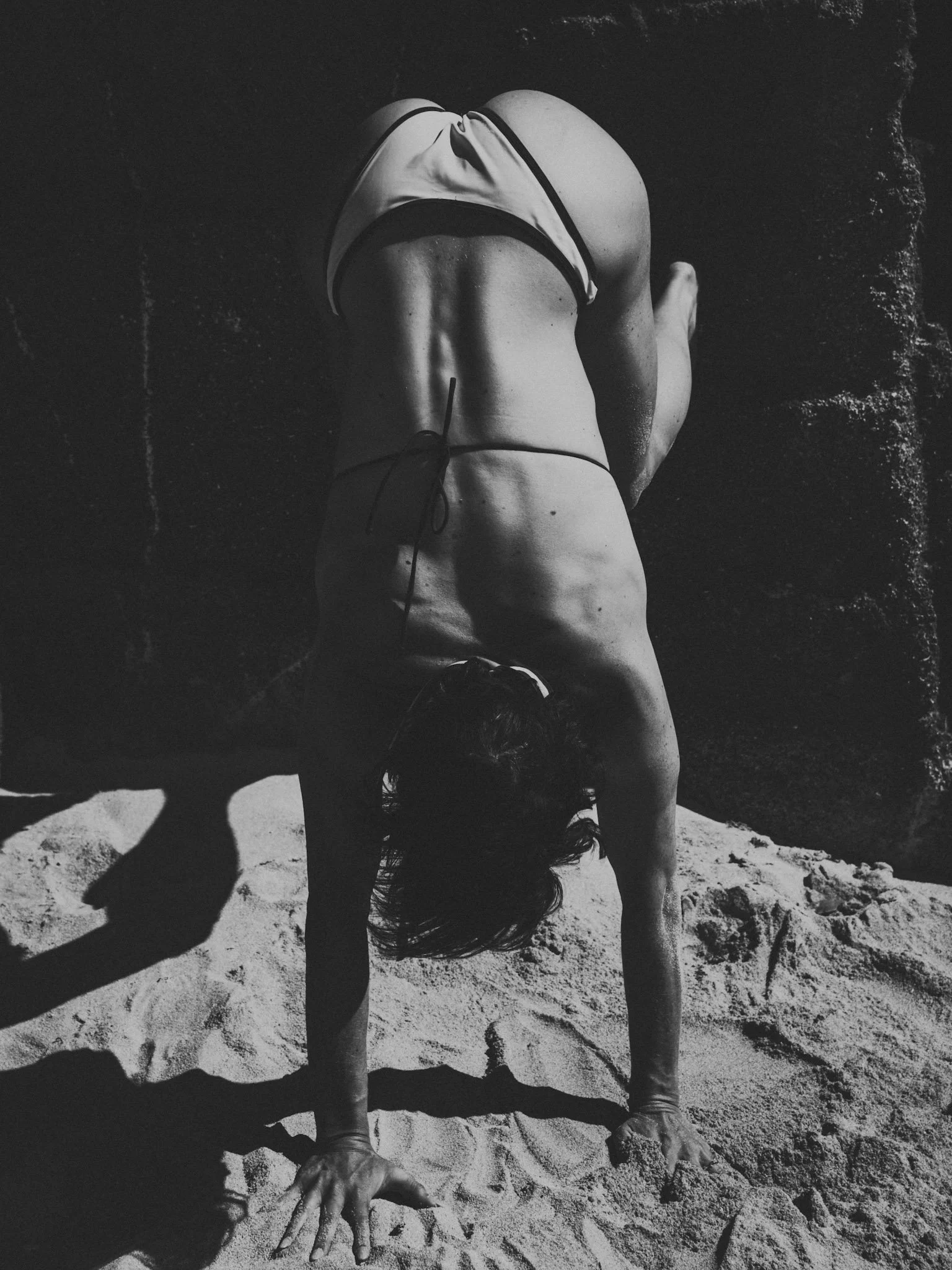 Schwarze und weiße Aufnahme einer Person, die eine Pflanzung macht, im Handstand auf Sand, mit einer Felsenwand im Hintergrund.