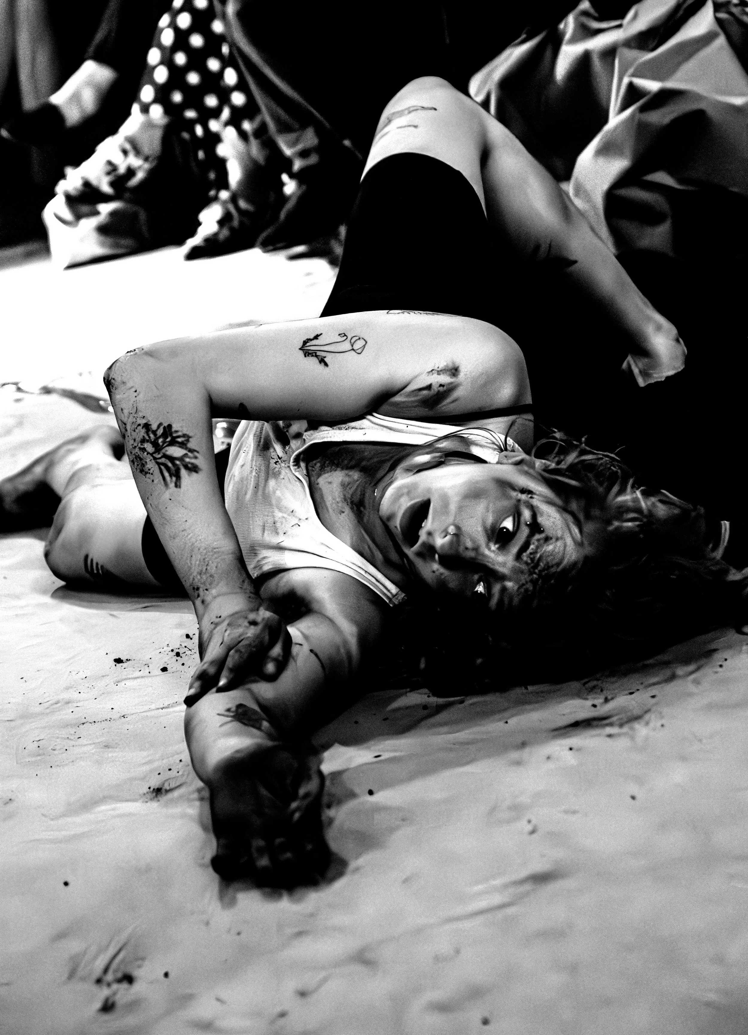 A black-and-white photo of a person in a white vest lying on the floor with an arm outstretched. On the floor around them is dirt.