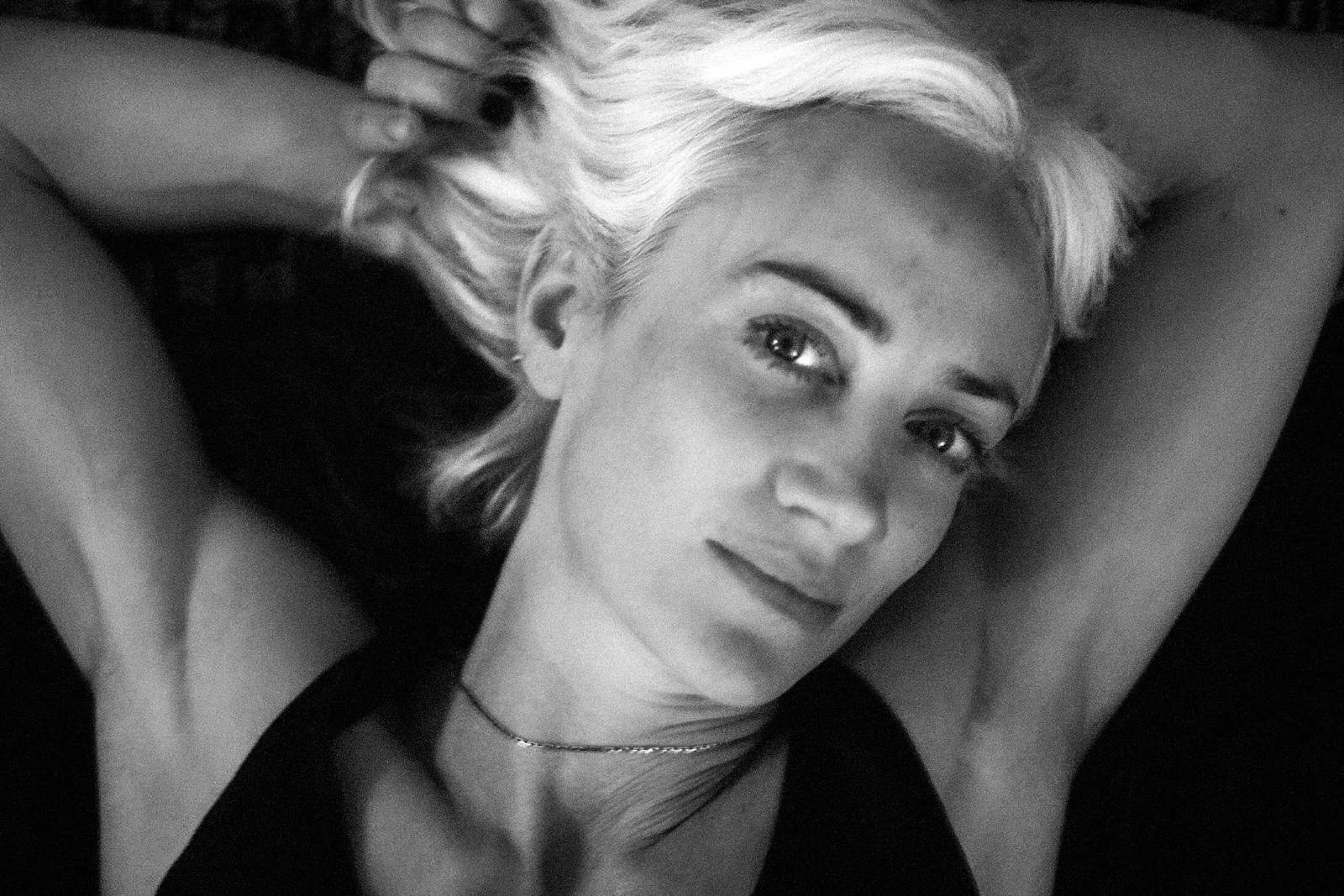 A black and white photo of a woman with blonde hair lying down with her arms behind her head, looking at the camera.