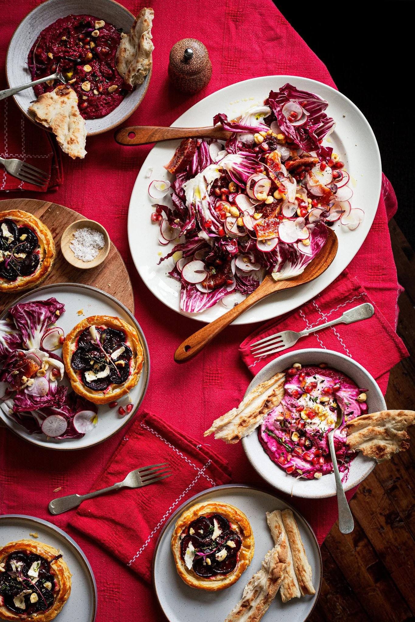 A table with various dishes including a large salad with radishes, pomegranate seeds, and endive, smaller bowls with beetroot dip and pitta bread, and individual plates with beetroot tartlets garnished with hazelnuts and herbs. Kathleen Gillan Photog
