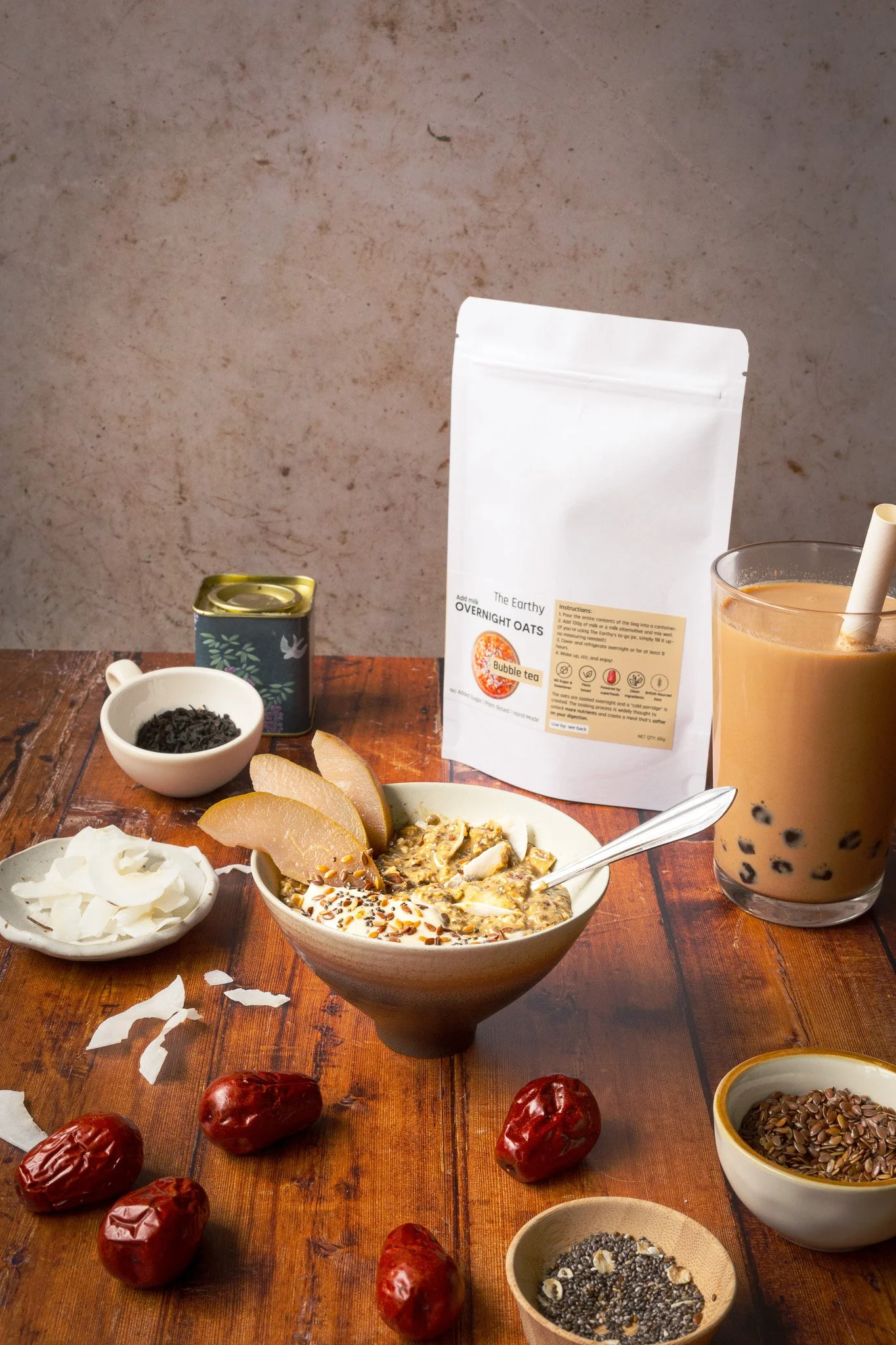 A bowl of The Earthy overnight oats topped with pear slices, seeds, and yogurt on a wooden table surrounded by dried jujube dates, dried coconut, flax seeds, chia seeds, and a glass of bubble tea with tapioca pearls. Kathleen Gillan Photography copyr