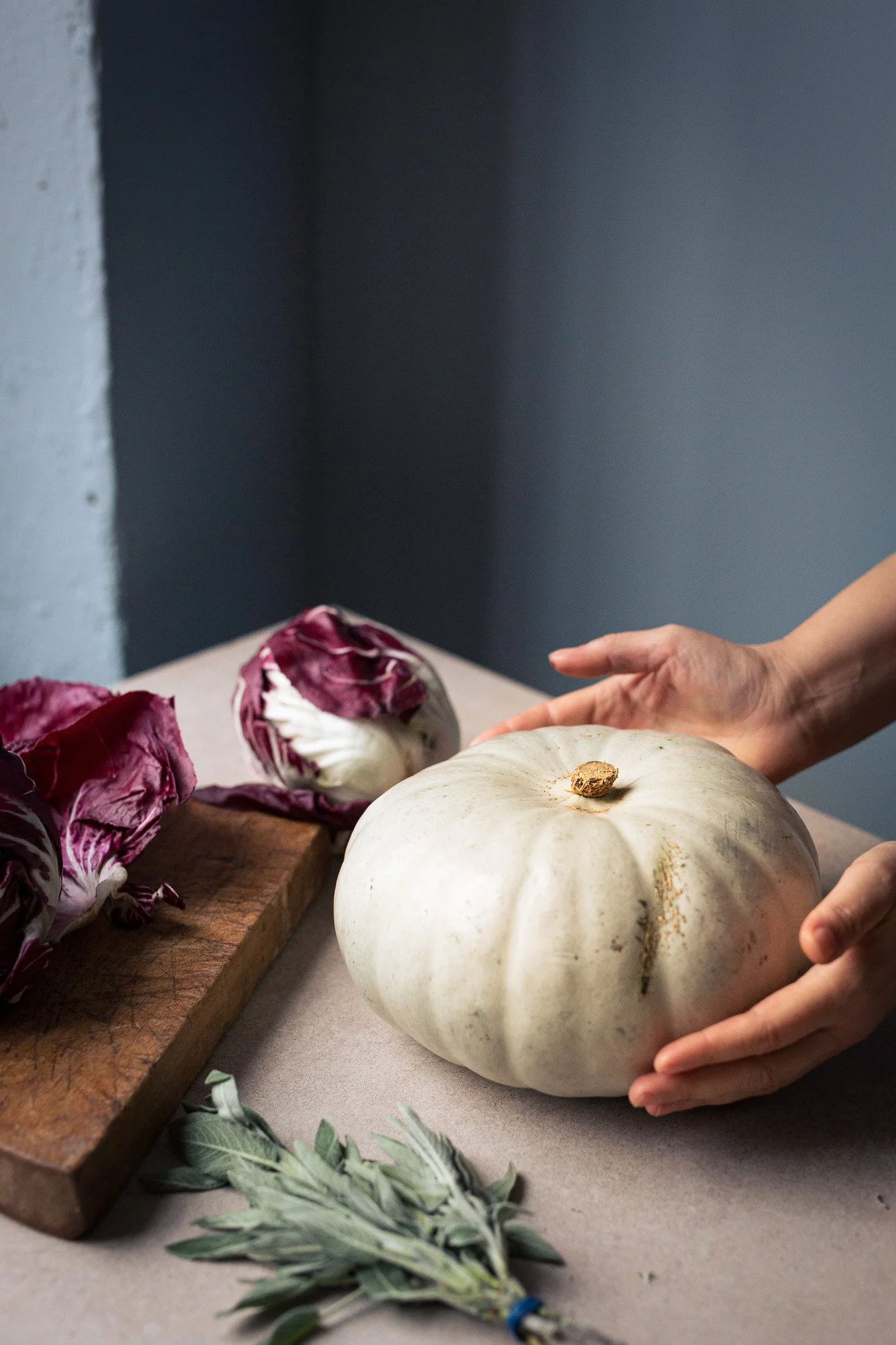 Person holding a large white pumpkin on a kitchen counter with purple radicchio, a sprig of sage, and a wooden cutting board. Kathleen Gillan Photography copyright-protected.