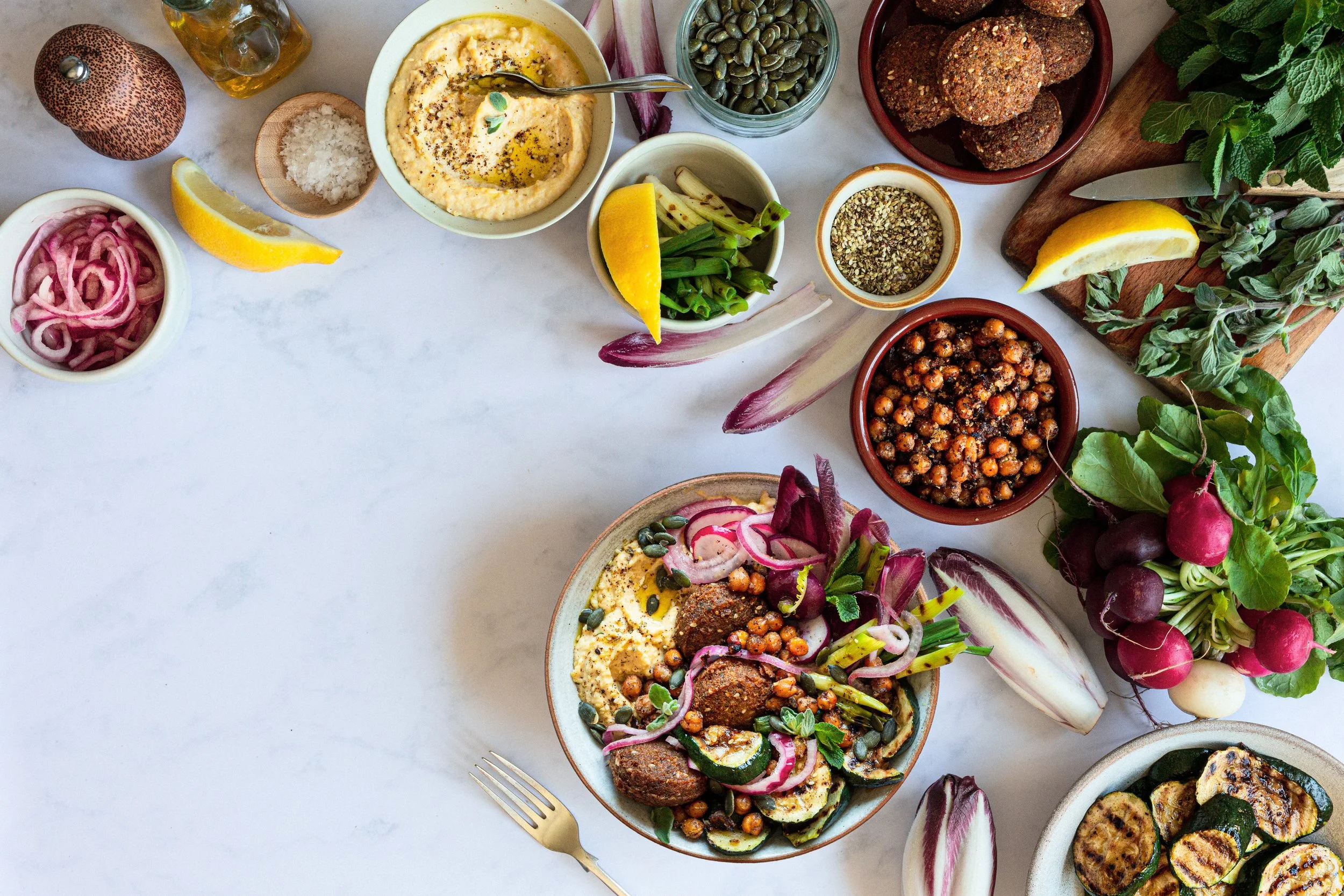 A spread of Mediterranean vegetarian dishes and fresh vegetables on a white surface, including hummus, grilled vegetables, chickpeas, radishes, cucumbers, herbs, lemon wedges, and condiments.