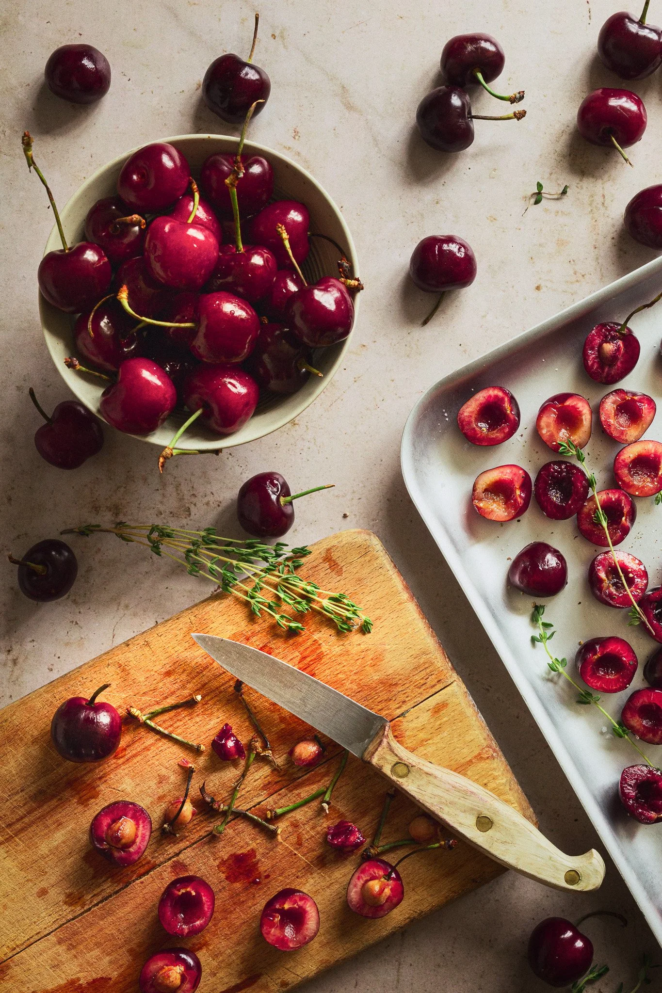 Fresh cherries on a wooden cutting board, with some halved cherries on a white tray, and a bowl of cherries on a countertop, with a knife and sprigs of thyme.