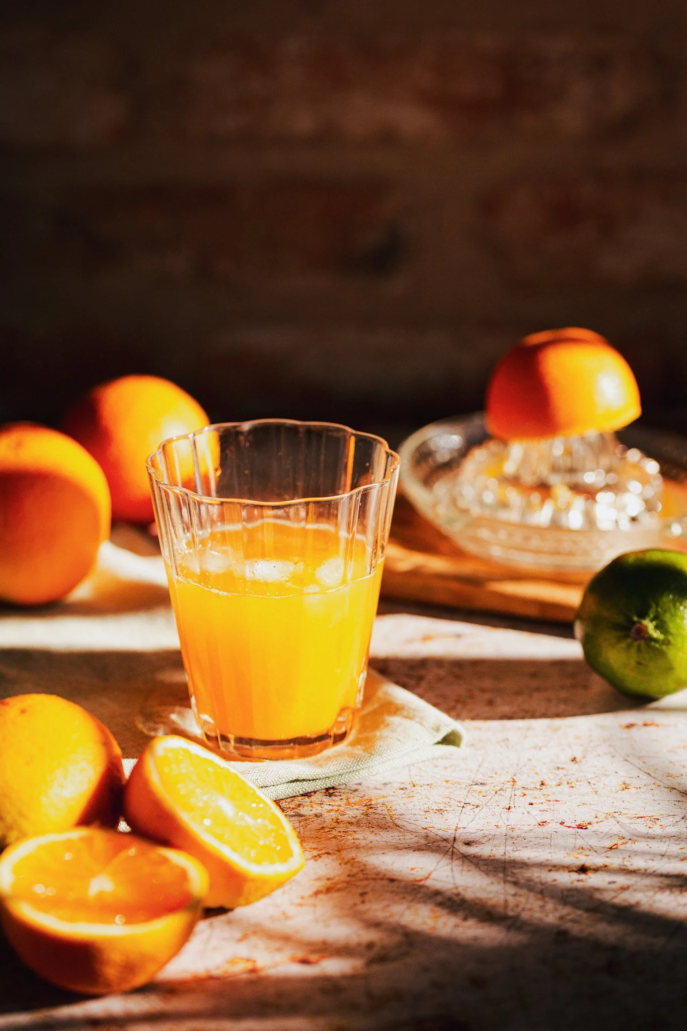 A glass of orange juice surrounded by whole and sliced oranges and a green lime, with a halved orange on a glass dish in the background.