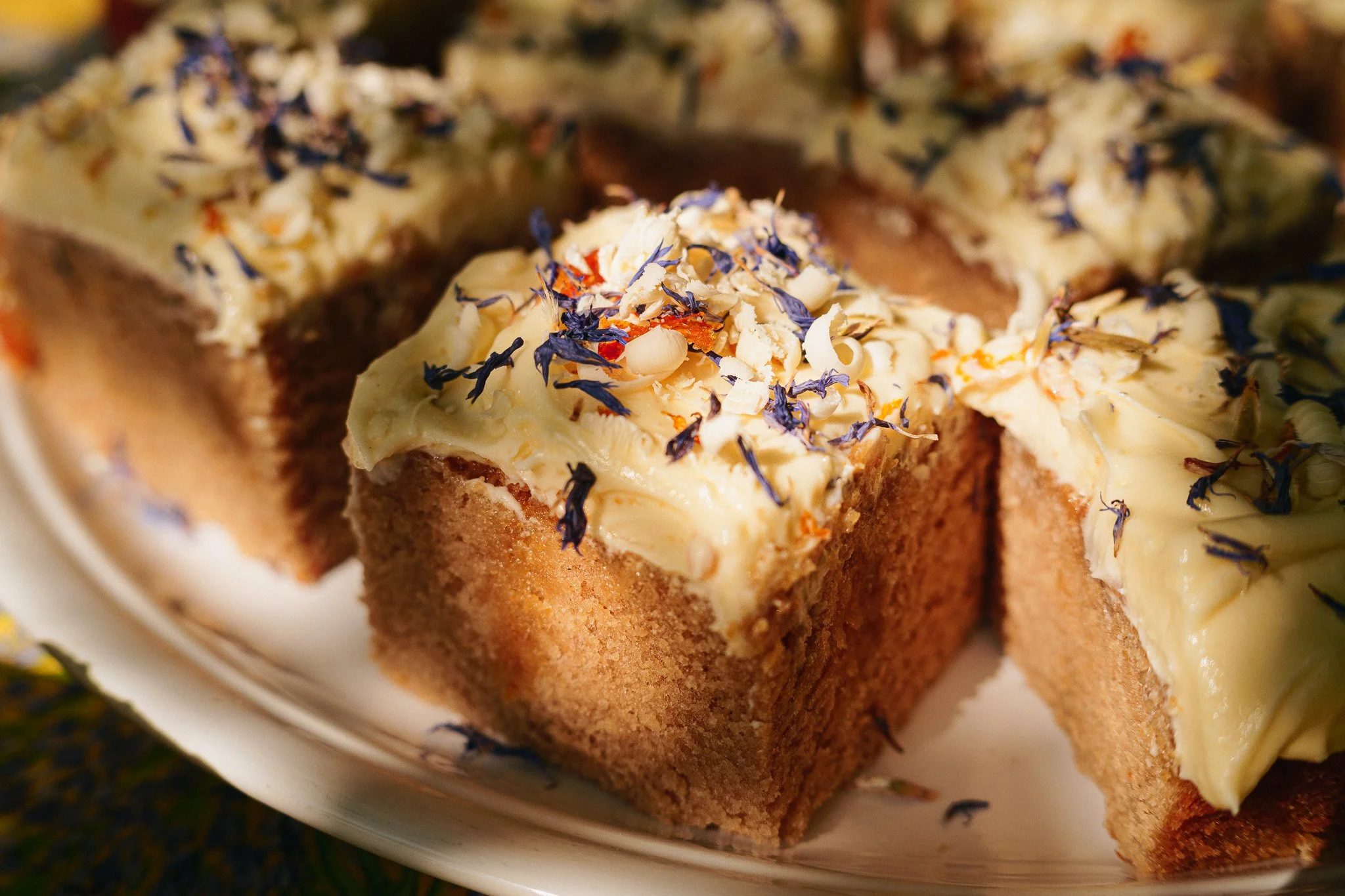 Close-up of a sliced cake with yellow frosting and edible flower petals on top, on a white platter.