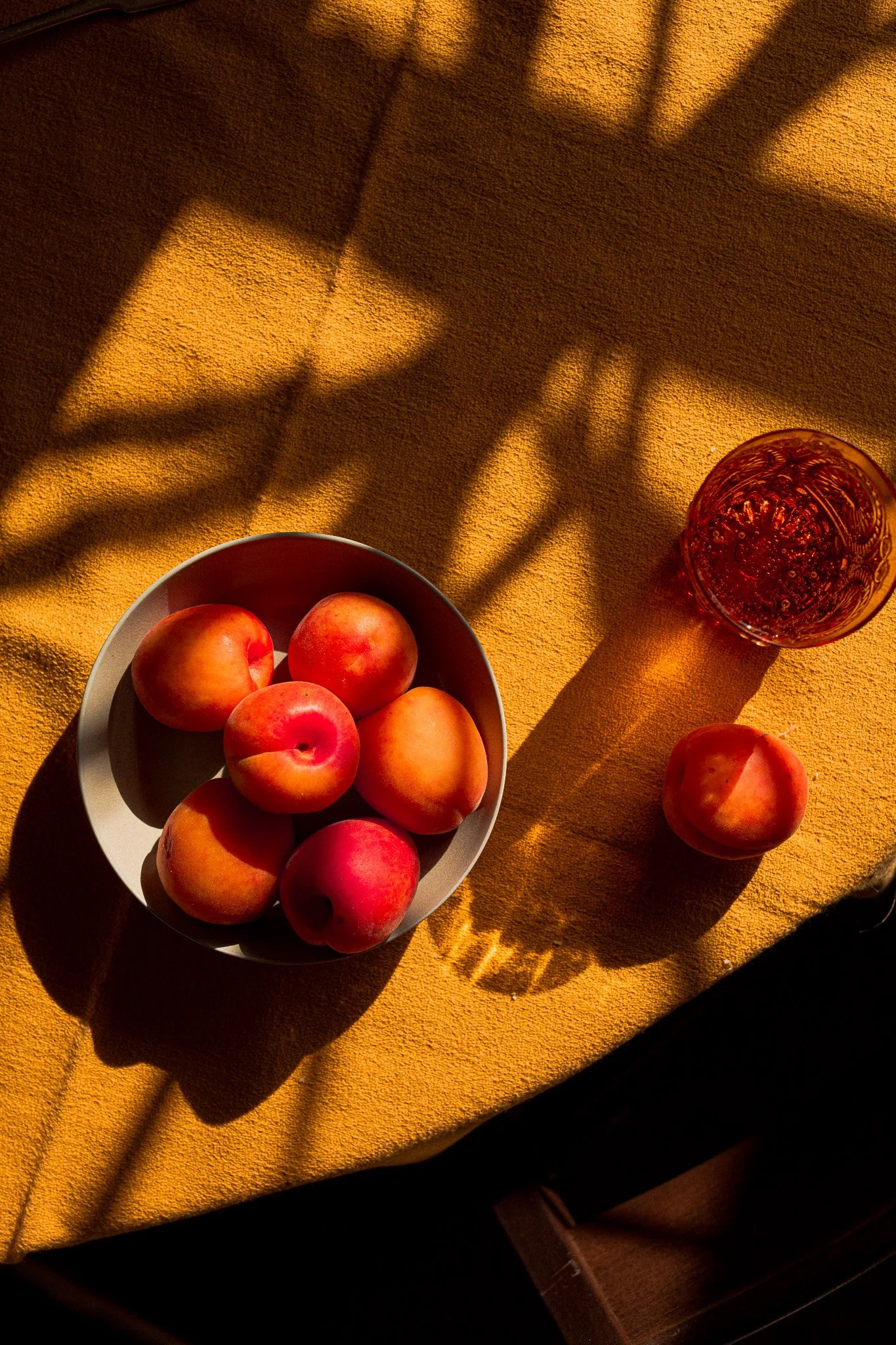 A bowl of six peaches on a yellow fabric surface with a glass of amber-colored beverage and a single peach beside it, all casting shadows in warm lighting.