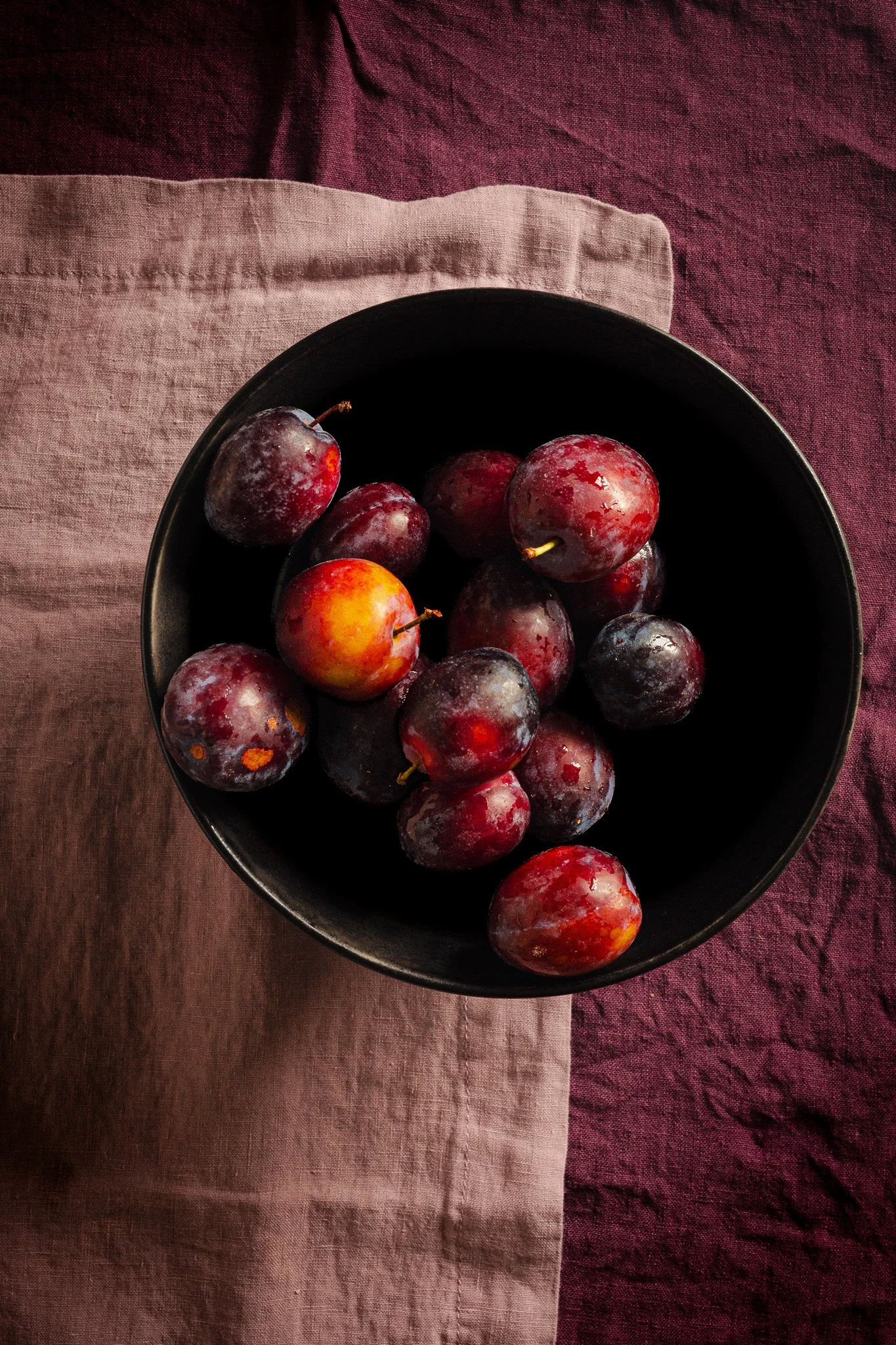 Black bowl filled with dark purple and red plums resting on two overlapping cloths, one beige and one deep red.