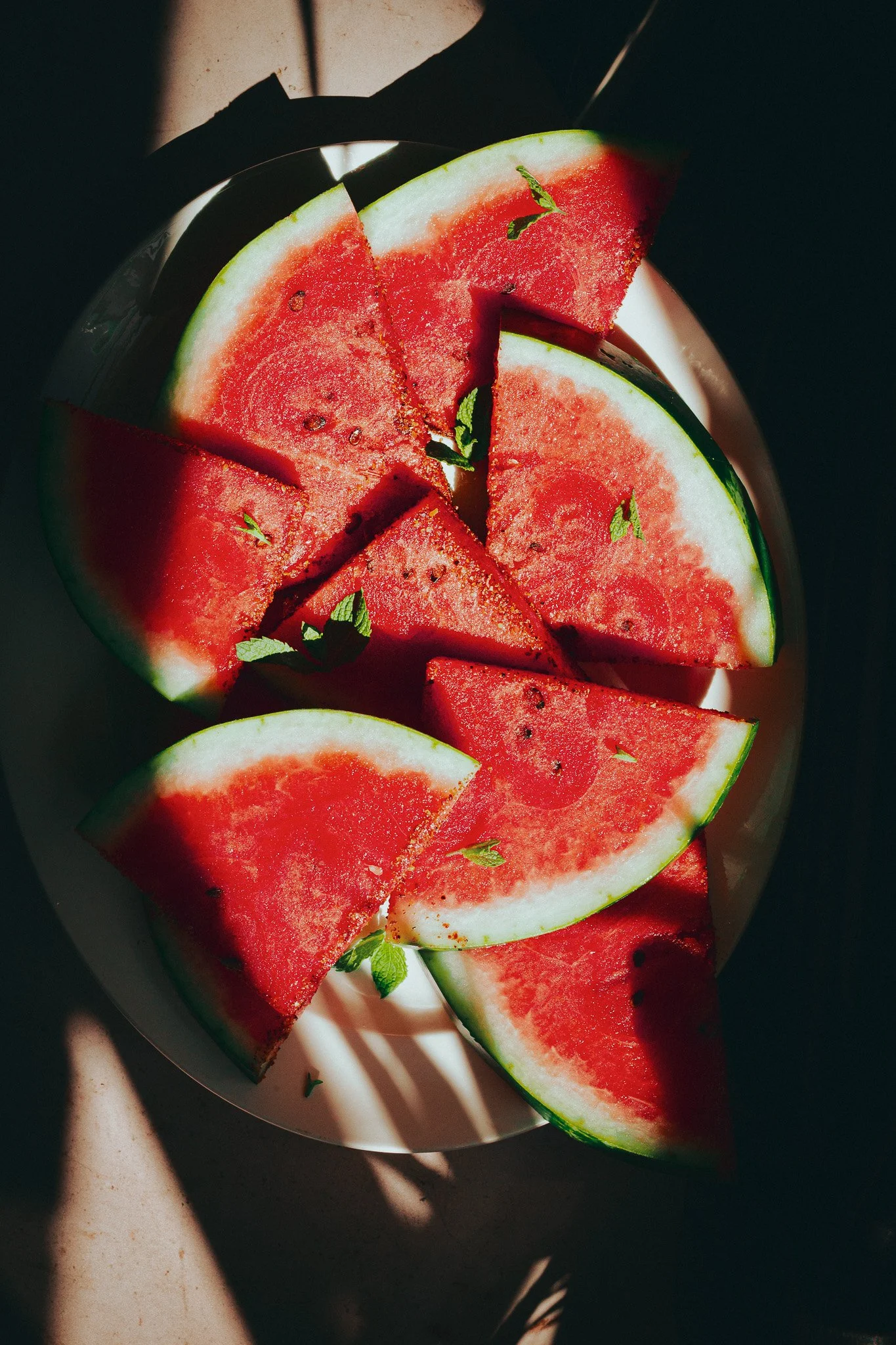 Slices of watermelon with mint leaves on a white plate, with shadows cast over the surface.