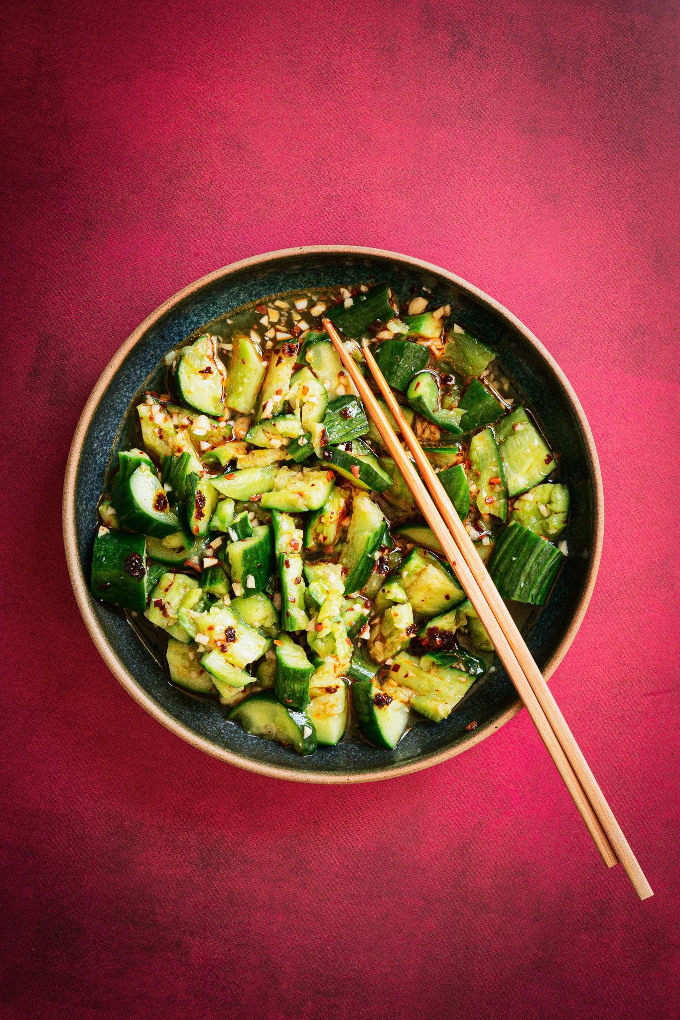 A bowl of chopped cucumbers with chili flakes and sauce, garnished with chopped garlic and green onions, placed on a red surface, with wooden chopsticks resting on top.