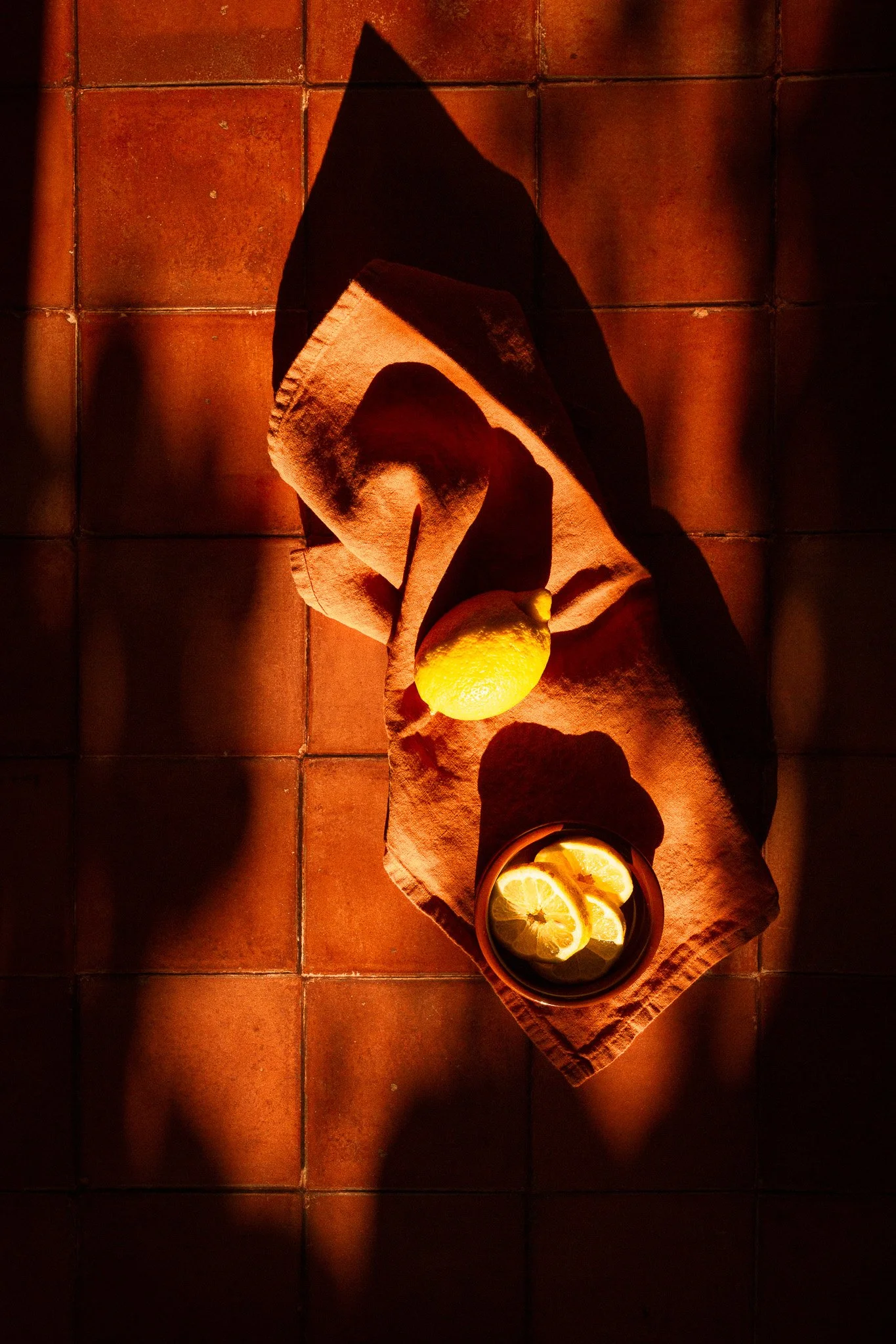 A lemon, a bowl of lemon slices, and a lemon on a textured cloth, arranged on a tiled surface with warm lighting and shadows.
