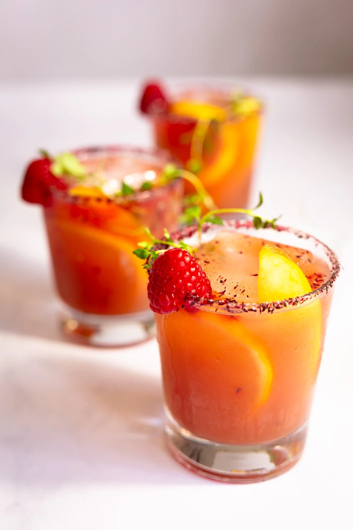 Three glasses of bright raspberry and orange juice garnished with raspberries, lemon slices, and green herbs on a white surface. Kathleen Gillan Photography copyright-protected.