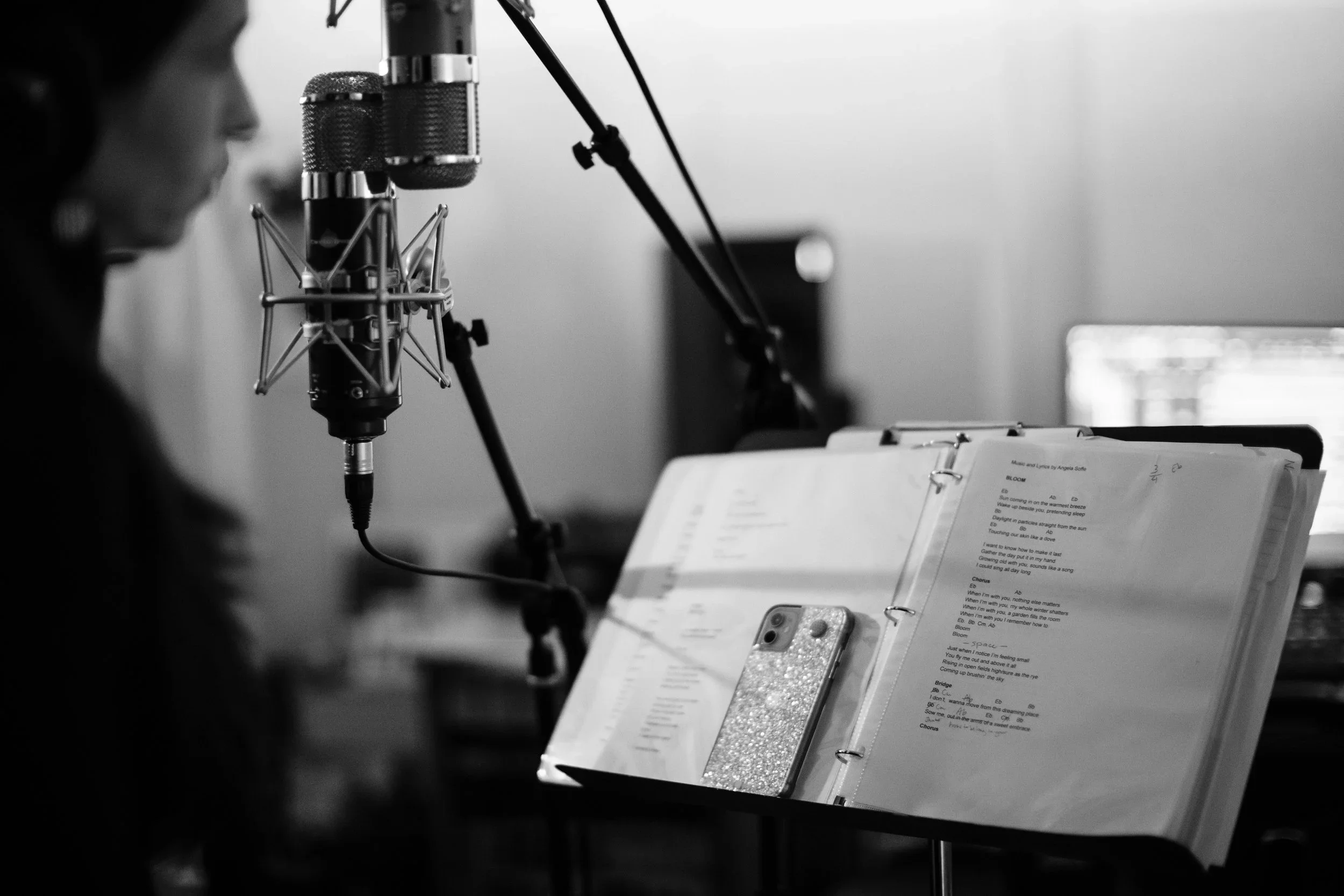 A woman recording vocals in a home studio with a microphone, music sheet stand, and a sparkly phone case.