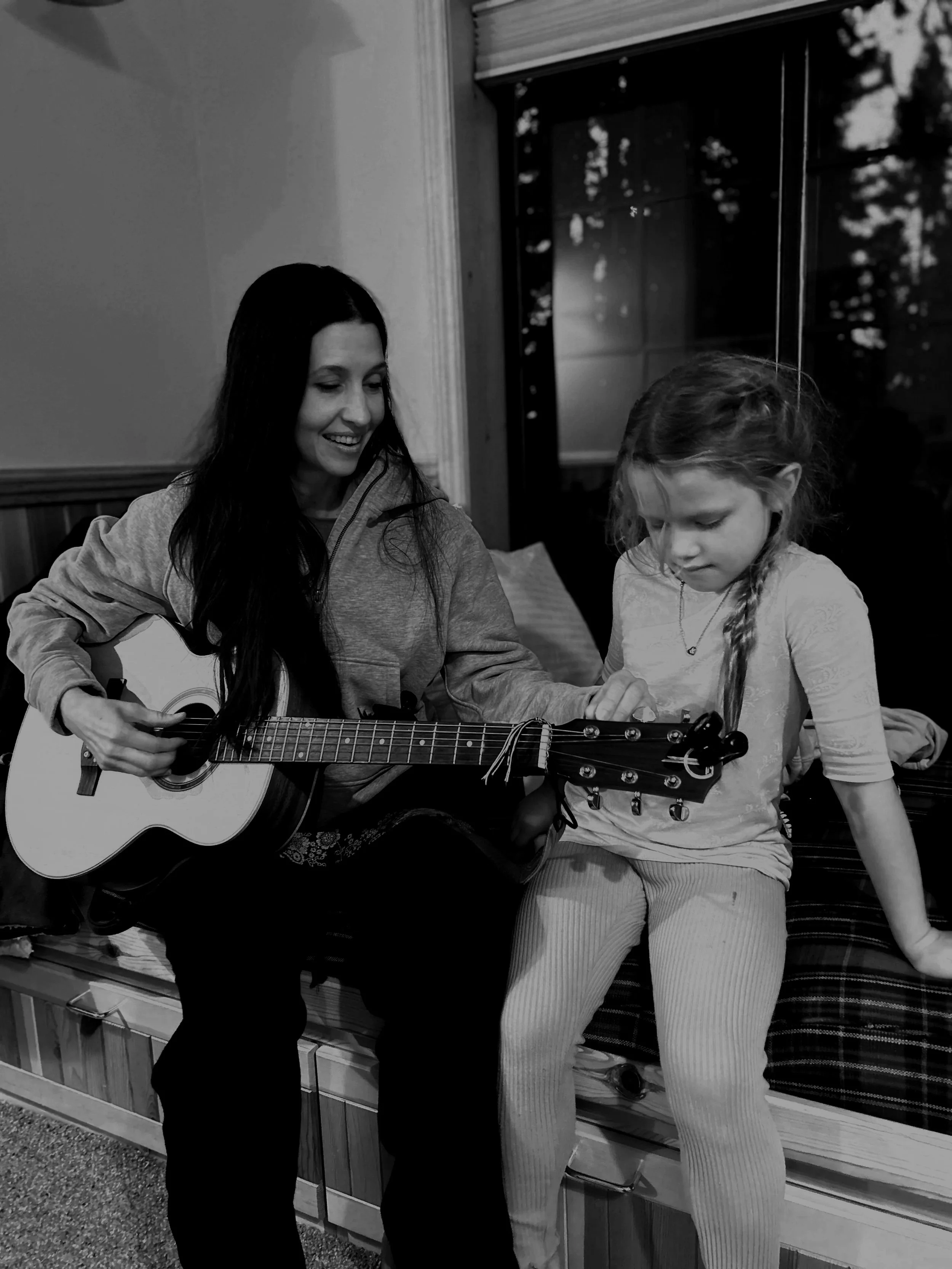 A woman with long dark hair playing guitar and smiling at a young girl sitting beside her, who is looking down. They are indoors near a window.