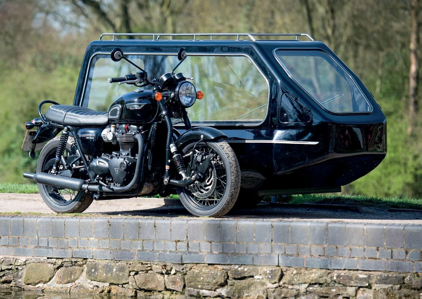 A black motorcycle with a sidecar sitting on a brick ledge by a body of water, with green trees in the background.