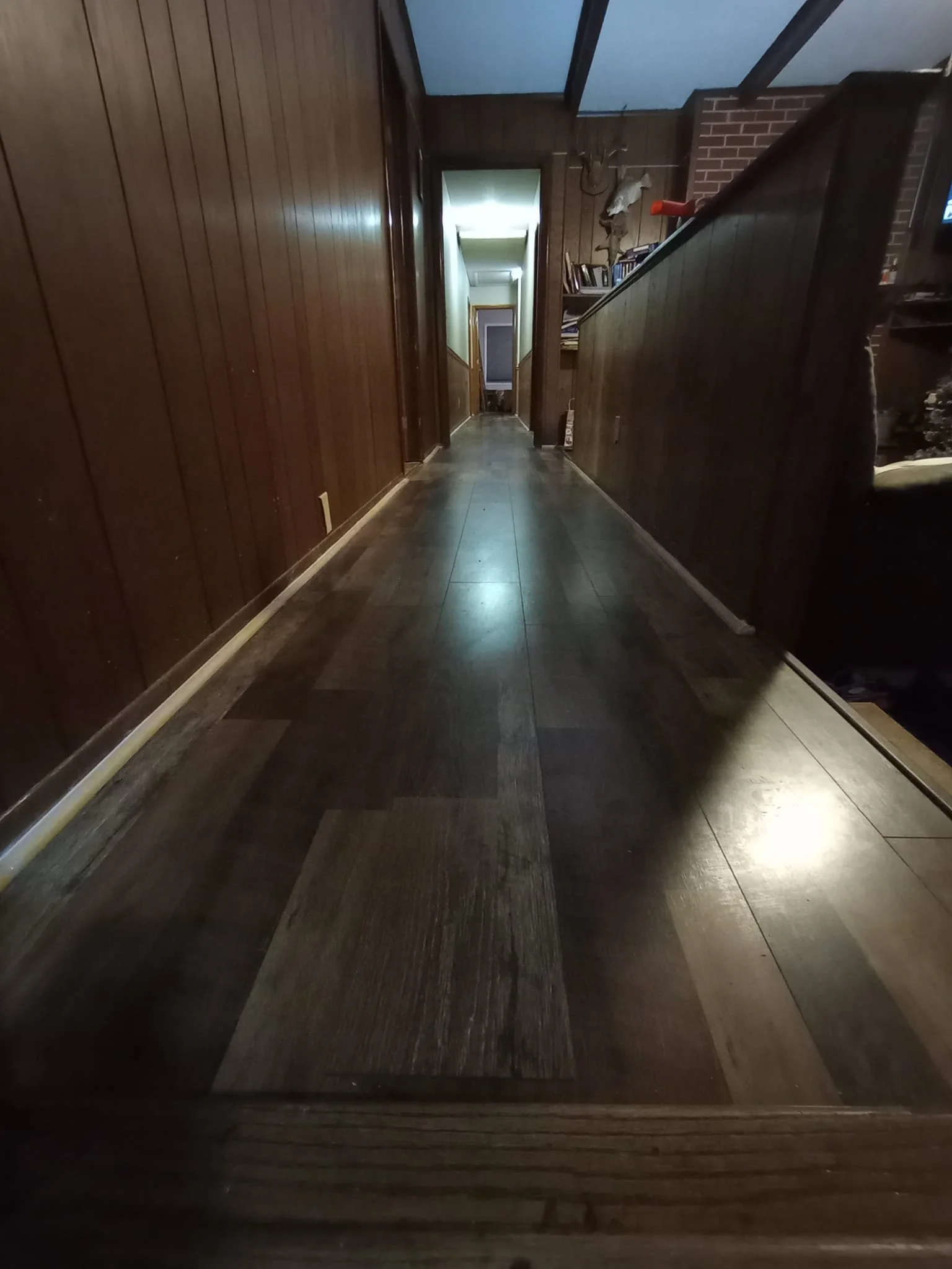 Long wooden hallway in a house with wood-paneled walls and dark hardwood floors, leading to a bright room at the end with a door.